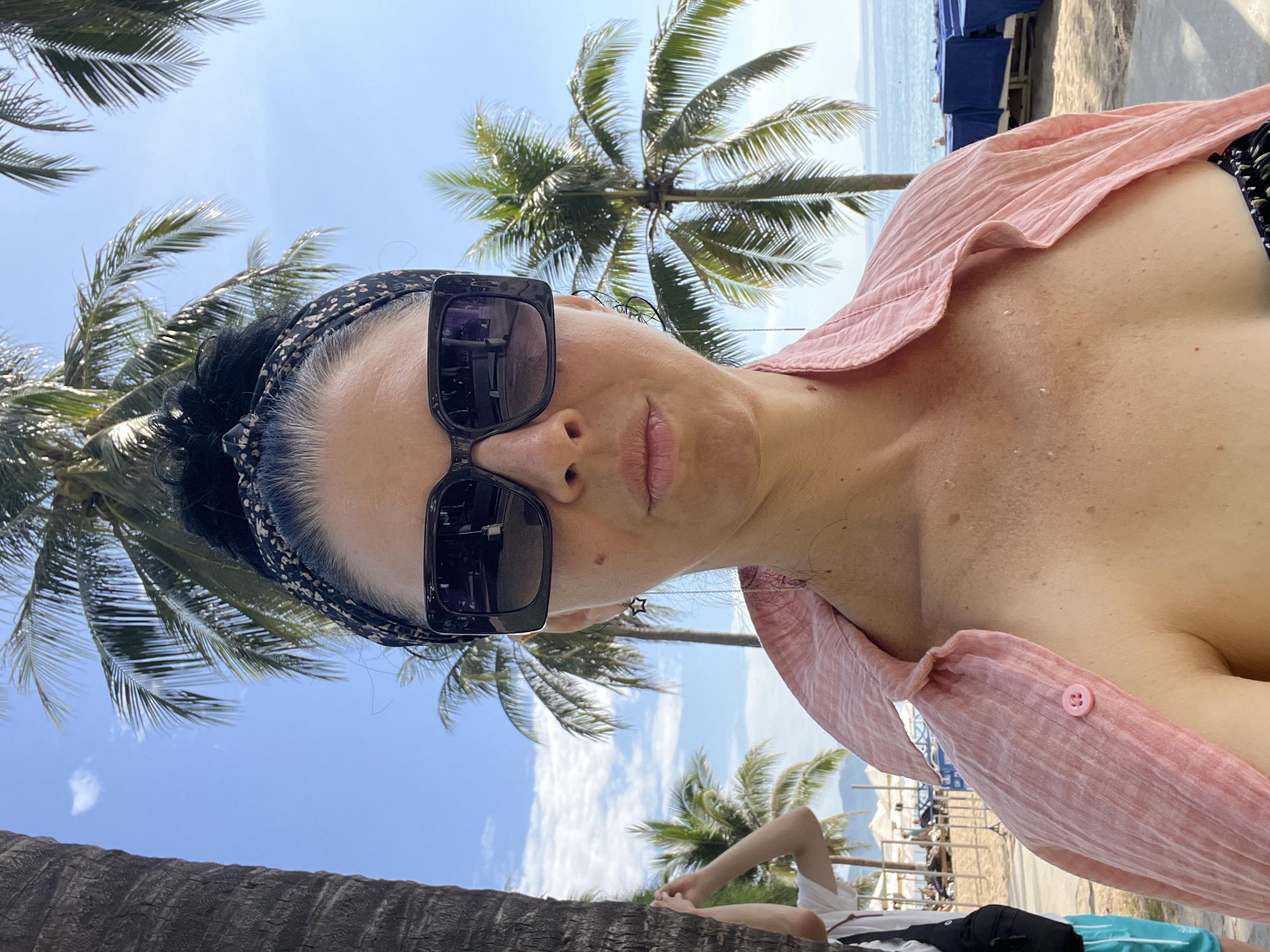 A woman is taking a selfie on a beach in Nha Trang, Vietnam. She is wearing a pink shirt, sunglasses, and a headband. Palm trees and the ocean are visible in the background. Another person is partially visible on the left side of the image.