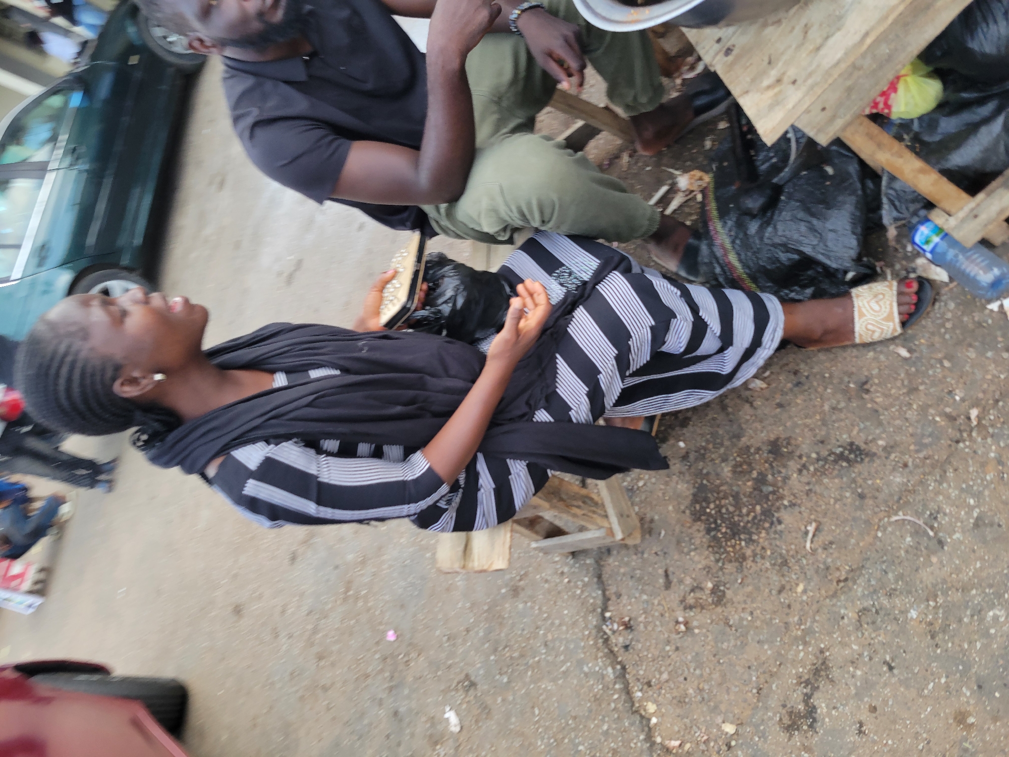 The image shows a scene in Jos, Nigeria, with several people and objects. In the foreground, a woman is seated, looking upwards with her mouth slightly open. She is wearing a black and white striped dress and a black headscarf. Her right foot is visible, adorned with a sandal featuring a heart design. Next to her is a black plastic bag. Behind her, a man is seated, wearing a black t-shirt and green trousers. He is looking down and appears to be engaged in a conversation or activity. Another person, partially visible, is also present.
The setting appears to be an outdoor area, possibly a roadside or market, with a rough, sandy ground. A green car is visible in the background, suggesting a public space. Wooden planks and a blue water bottle are also present in the scene, hinting at informal commerce or daily life activities. The overall lighting suggests it could be daytime. The expressions of the individuals are somewhat neutral, but the woman's open mouth might indicate surprise, speech, or exertion. No visible text is present in the image.