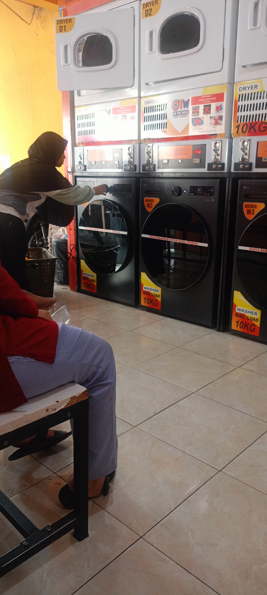 The image shows a laundromat in Mekar Rahayu, Indonesia. In the foreground, a person wearing a red top and light blue pants is sitting on a bench. To the left of the person is a black laundry basket. Behind the bench are several washing machines and dryers. A woman wearing a black hijab and a gray and black top is operating one of the washing machines. The walls are painted yellow, and the floor is tiled. The washing machines are labeled with "WASHER MAX. LOAD 10KG" and "DRYER". One of the dryers is labeled "DRYER D1" and another "DRYER D2". Another dryer is labeled "DRYER 10KG". The overall scene depicts a functional laundromat with customers present.