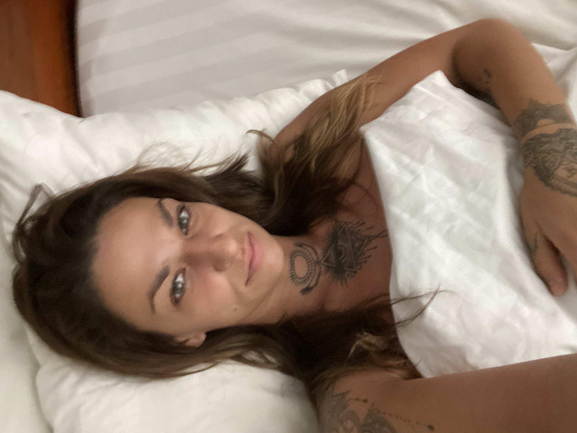 A woman is lying in a bed covered with white sheets. She has shoulder-length brown hair and light skin. A tattoo is visible on her neck and another on her arm. Part of a wooden headboard is visible in the background. The image was likely taken indoors, but the specific location in Thủ Đức, Vietnam, cannot be determined from the image.