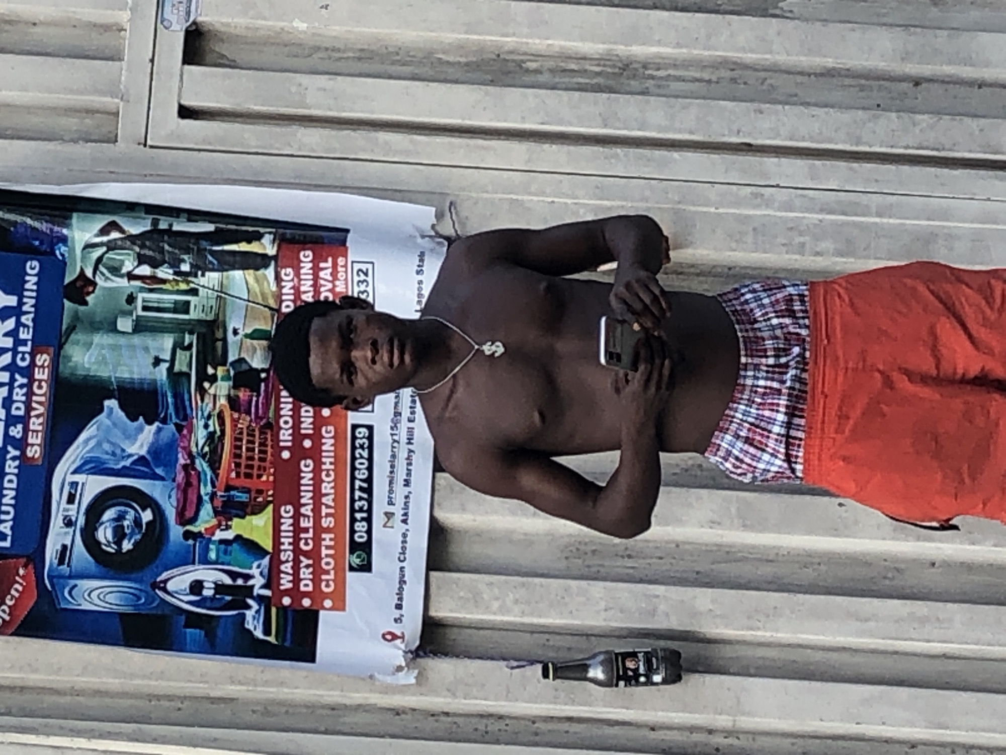 A shirtless man lies on a concrete structure in what appears to be a public space. He is holding a smartphone. He wears plaid underwear and red shorts, and a dollar sign necklace. Next to him is a laundry service poster with a phone number and an address in Balogun Close, Akins, Marshy Hill Estate, Lagos State, Nigeria. A small bottle lies near the poster. The concrete structure has a series of horizontal grooves.
