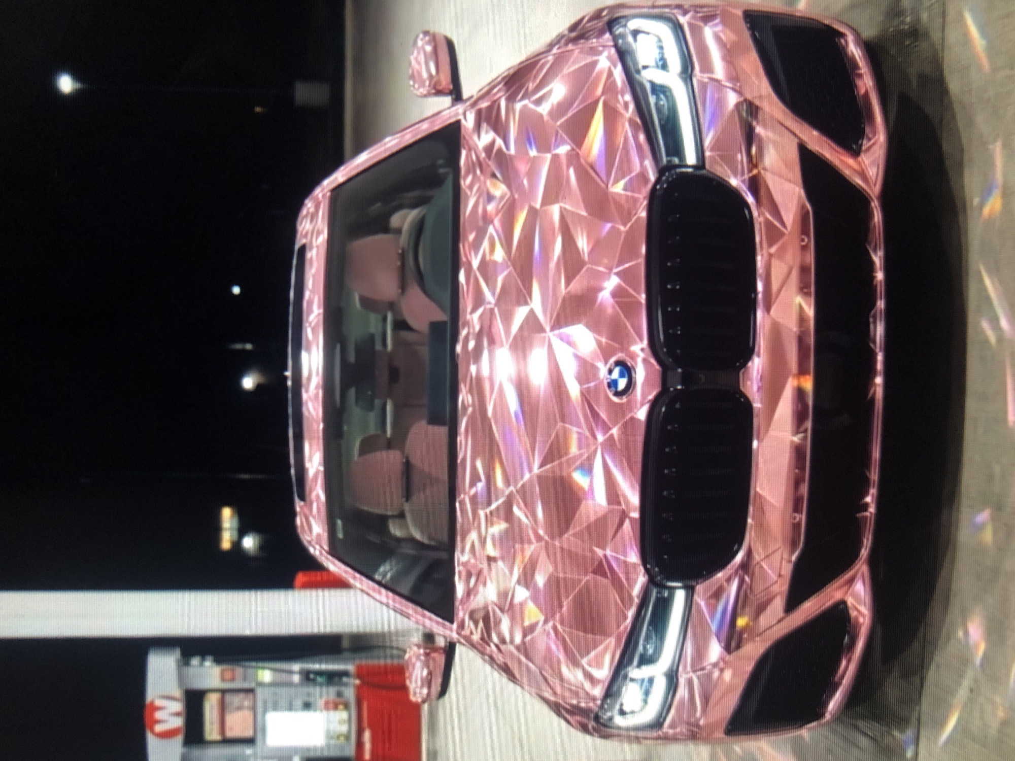 A pink BMW with a geometric, diamond-like pattern is parked at a gas station with a red "W" sign. The scene appears to be at night, with streetlights visible in the background. The location is not specified, and I cannot locate this in Lhokseumawe, Indonesia. The car's headlights are on.