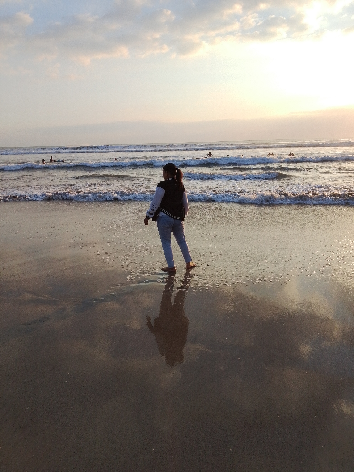 A full shot captures a person standing on a wet, sandy beach in Kuta, Indonesia, facing the ocean. The individual, seen from behind, wears a black and white bomber jacket and light-colored pants. Their shadow is clearly visible on the wet sand in front of them. In the distance, several people are in the water, some appearing to be swimming or surfing. Gentle waves are breaking on the shore, and the sky above is a soft gradient of pale blue and orange, indicating either sunrise or sunset. The overall scene is tranquil, with the reflective wet sand mirroring the sky and the faint outlines of people in the ocean.