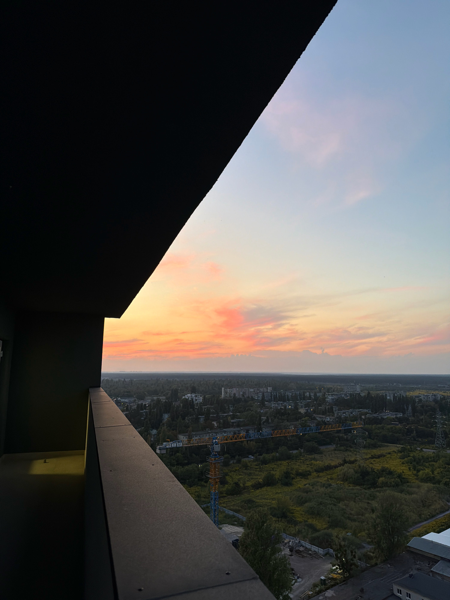 The image depicts a view from a high-rise building's balcony in Kyiv, Ukraine, overlooking a cityscape at sunset. The foreground features a dark, angular balcony railing and wall, casting a shadow. The midground reveals a sprawling urban landscape dotted with trees and buildings, including a prominent construction crane with yellow and blue accents. The background is dominated by a vibrant sunset sky with hues of orange, pink, and pale blue, with thin clouds scattered across. The overall impression is of a serene evening over an active urban area.