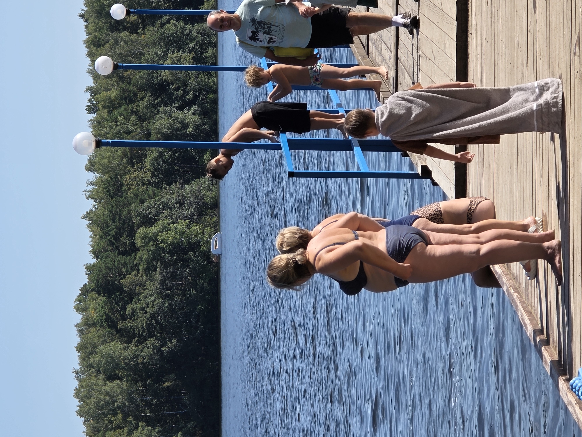 A group of people are gathered on a wooden pier on a sunny day, with a lake and a forested shoreline in the background. The pier has blue railings and lampposts. In the foreground, two women in swimsuits are standing, facing the water. Behind them, a boy is leaning over the railing. Further down the pier, a man in a t-shirt and shorts is walking, carrying a bag. Two young boys are also on the pier, one in swim trunks and the other wearing a towel like a cape. A white inflatable object is visible in the lake. The overall scene suggests a recreational outing by a lake, likely during the summer months. The location is Chebarkul, Russia.