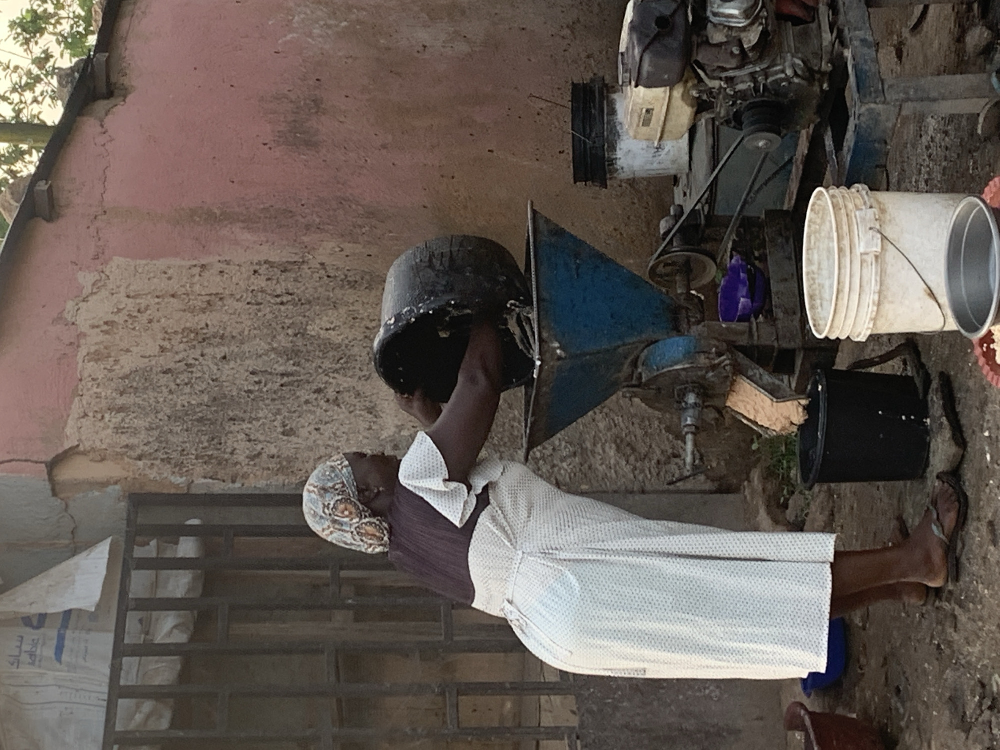 In the city of Jos, Nigeria, a woman is shown from the side, wearing a headscarf and a long, light-colored dress. She is leaning over a blue and metal processing machine, holding a large black container, seemingly to pour its contents into the machine. To the right of the woman and the machine, there is a small engine, possibly for powering the processing unit. Various buckets and containers are present around the scene, some empty and others appearing to hold processed material. The environment suggests an outdoor, possibly domestic or small-scale industrial setting, with weathered walls and a dirt or gravel ground. The overall scene depicts a moment of manual labor, likely related to food processing or a similar agricultural activity.