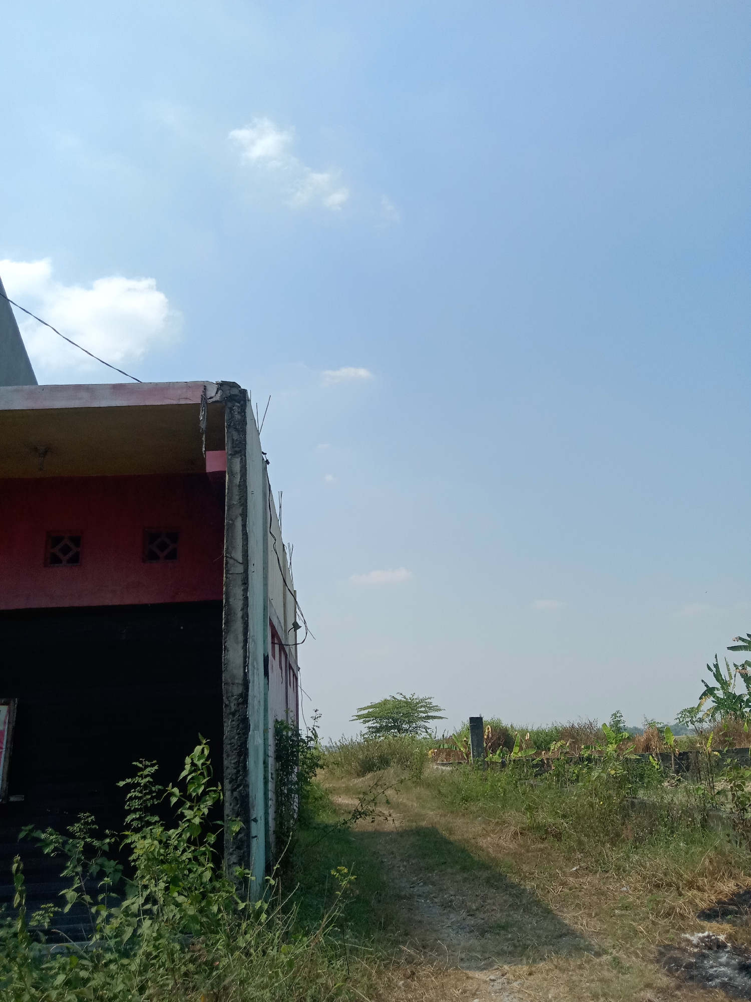 The image captures an outdoor scene in Kemamang, Indonesia, under a clear blue sky with scattered white clouds. In the foreground on the left, the side of a partially constructed or dilapidated building is visible. It has a dark lower section, a red middle section with two square decorative vents, and a lighter upper section with exposed concrete and rebar at the edge, suggesting unfinished construction or decay. Electrical wires are strung across the top of the building.
A dirt path leads away from the building into a grassy and vegetated area. The path curves slightly to the right. Along the path and to the right of it, there is abundant vegetation, including tall grasses, bushes, and some banana trees in the background. In the mid-ground, there is a single, small, leafy tree. Further in the distance, the landscape appears to be rural or semi-rural, with more vegetation and possibly other structures or remnants of construction. The overall impression is one of a somewhat neglected or undeveloped area. There are no people or animals visible in the image.