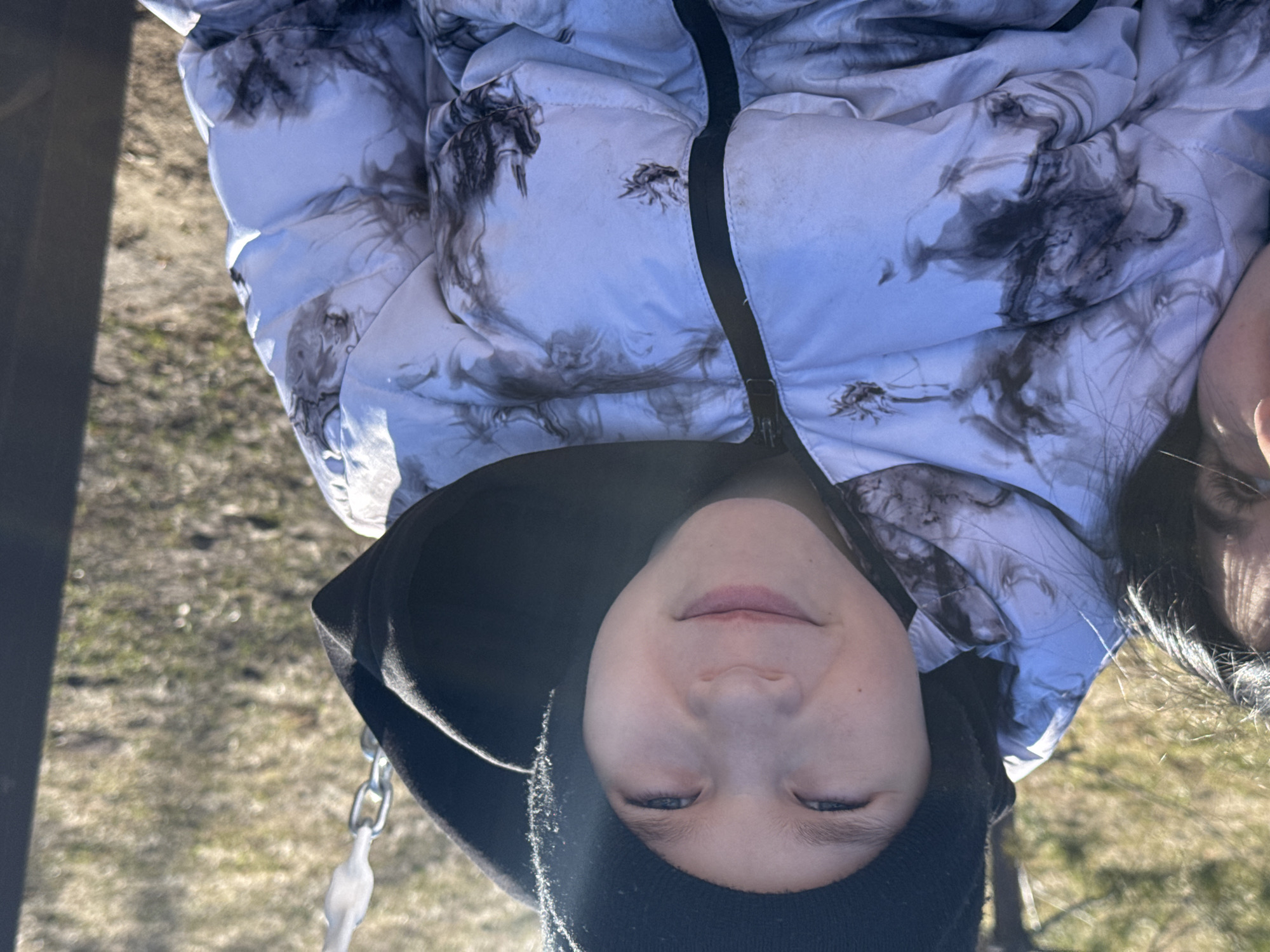 A child wearing a black hat and a white puffy jacket with gray and black patterns is upside down. A swing chain is visible. The location is not specified in the provided data.
