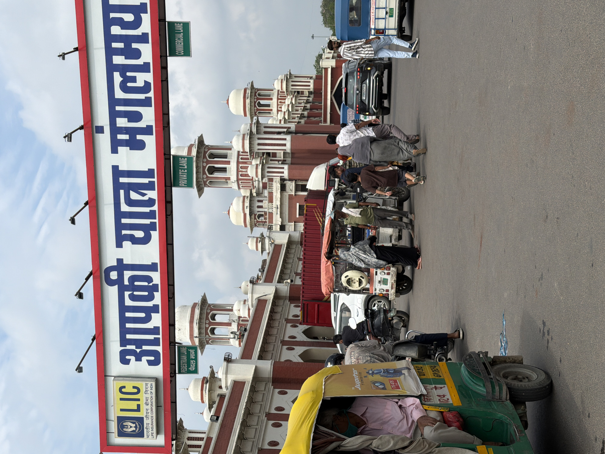 The image shows a street scene in front of a red and white building with multiple domes and arches, characteristic of Indian architecture. A large banner with the text "LIC आपकी यात्रा मंगलमय" (LIC Your journey is auspicious) is prominently displayed overhead, indicating the presence of the Life Insurance Corporation of India. Several vehicles, including auto-rickshaws, a car, and a van, are on the road. Pedestrians are walking in various directions. Signs for "PEDESTRIAN LANE," "PRIVATE LANE," and "COMMERCIAL LANE" are visible, suggesting traffic management within the area. The overall environment appears to be a busy public space in the city of Lucknow, India.