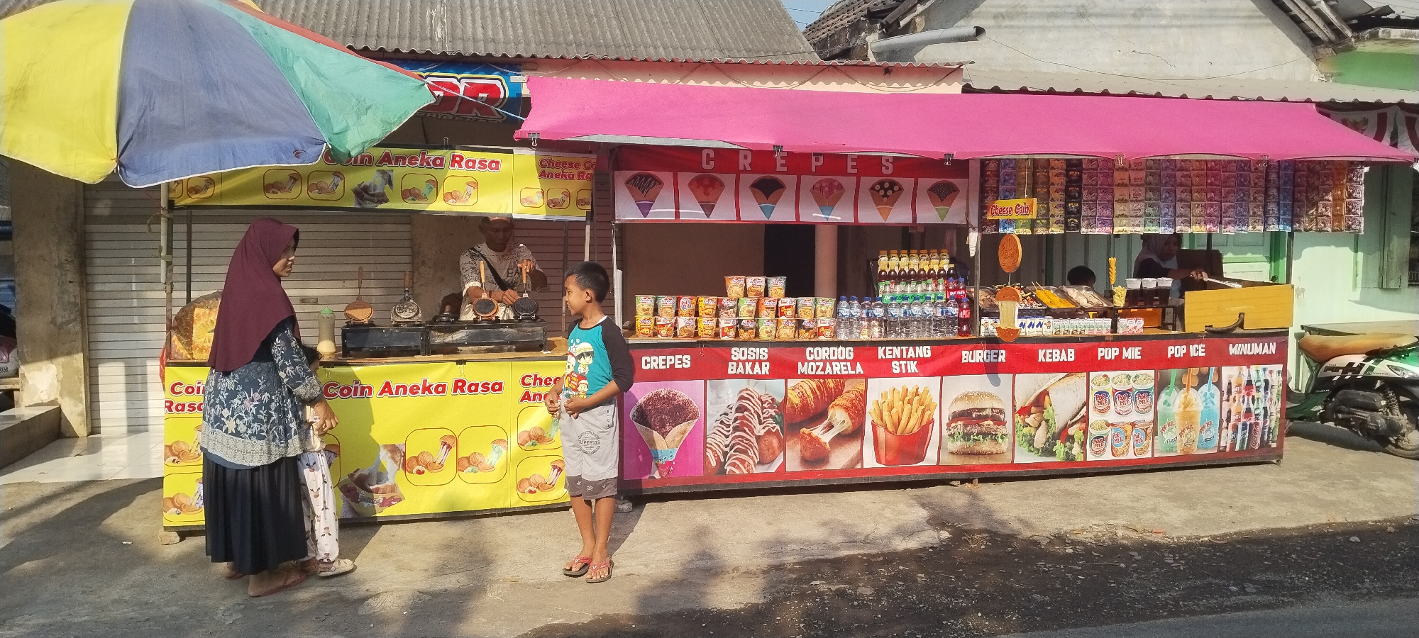 In Yosowilangun, Indonesia, a street food stall is set up with a colorful awning and a variety of food items displayed. The stall features two distinct sections: one selling "Coin Aneka Rasa" (various flavors of coin cakes) and another offering crepes, sausages, mozzarella hot dogs, french fries, burgers, kebabs, instant noodles, ice pops, and drinks.
A man, presumably the vendor, is seen working at a waffle maker in the "Coin Aneka Rasa" section. A woman wearing a hijab and a patterned dress, accompanied by a young child in patterned pants, stands near the stall, possibly as customers or family. A young boy in a blue and black t-shirt and grey shorts stands facing the crepe section, looking at the food displays.
The stall is decorated with images of the food items it sells. Behind the stall, the background shows a closed rolling door of a building, and further back, a residential house with a corrugated metal roof. A motorcycle is parked on the right side of the frame, partially obscuring the end of the food stall. The ground in front of the stall is paved with concrete. The scene is lit by natural sunlight, casting shadows on the ground.