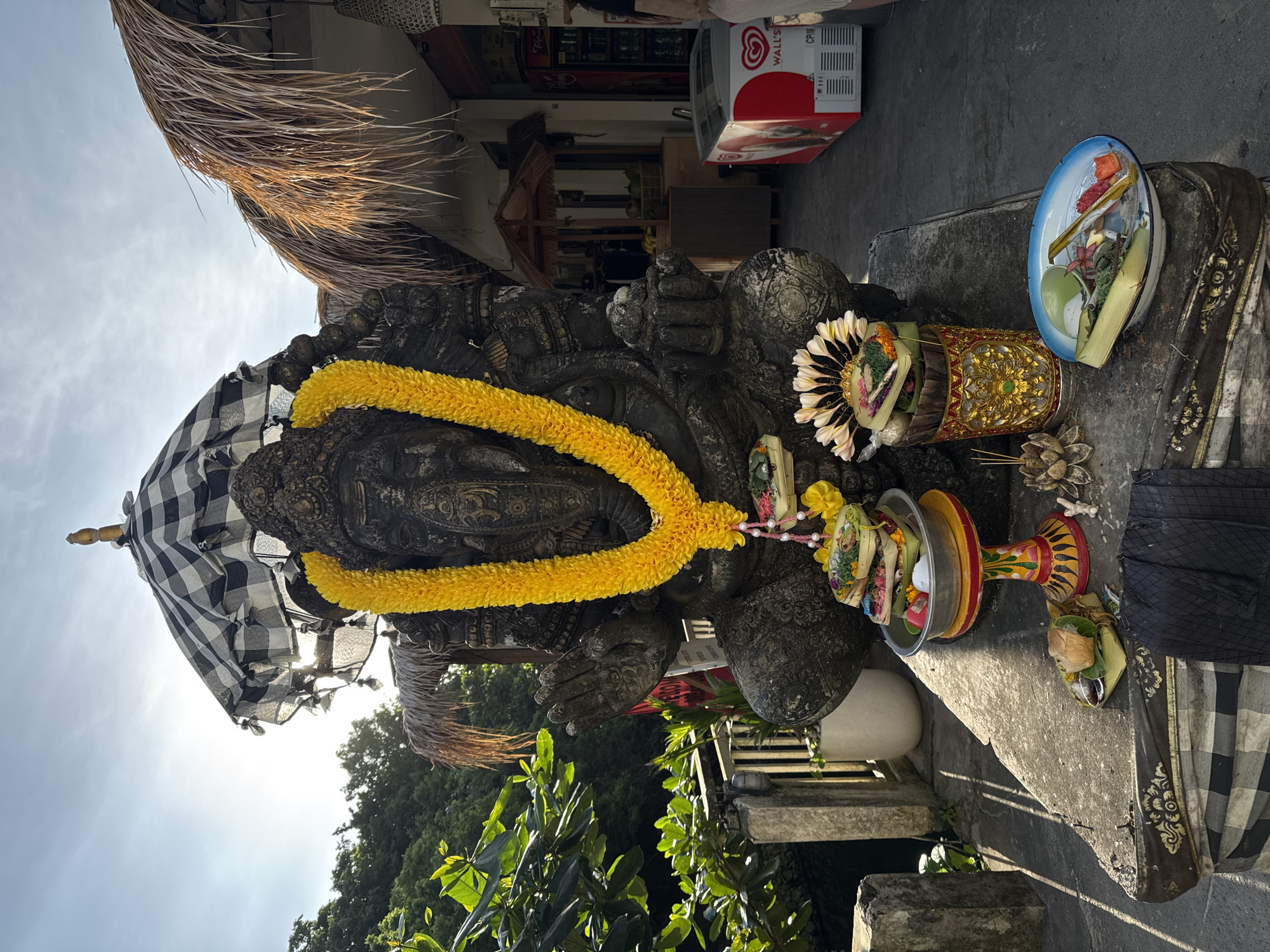 The image shows a Balinese statue adorned with a yellow garland and a checkered cloth, likely a sacred object or deity, set on a stone platform. Offerings of flowers, fruits, and other items are arranged around the statue. The statue appears to be located in an outdoor setting, possibly a temple or public space in Kuta Selatan, Indonesia, as suggested by the Balinese cultural elements. The background includes greenery and buildings, with a beverage cooler visible, indicating a mix of traditional and modern elements. The shot seems to be taken from a low angle, with the sky visible above.