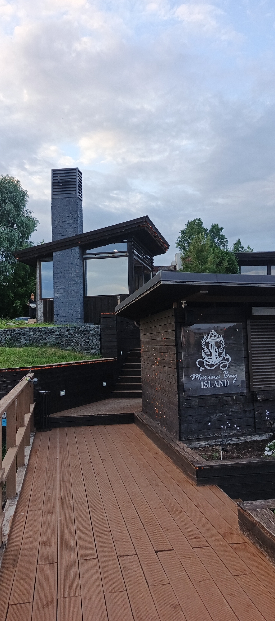 This is a photograph of a modern wooden building with a large stone chimney, taken outdoors in Votkinsk, Russia. The building features dark wooden siding, a sloping dark roof, and large windows that reflect the sky. A wooden deck and railing are in the foreground, leading towards the building. A sign with an anchor logo and the words "Marina Bay ISLAND" is affixed to one of the wooden structures. A person is visible standing on a stone retaining wall to the left of the main building. The sky is overcast with clouds. The setting appears to be a waterfront or waterside location, likely a restaurant or resort.
