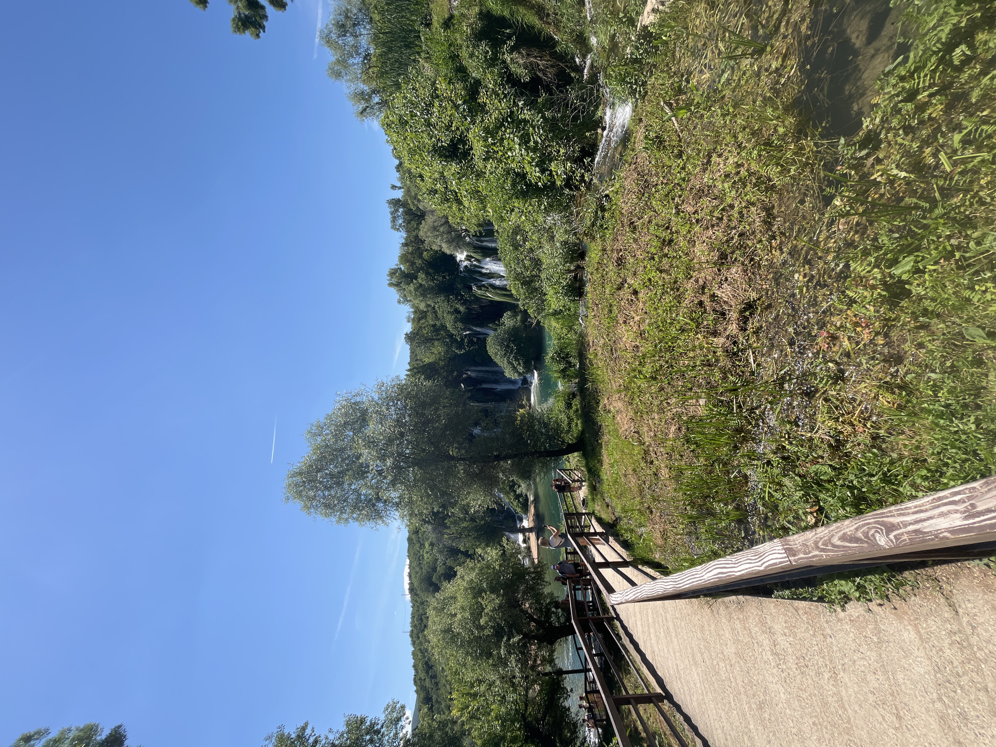 The image depicts a scene at Kravice Falls, located in Bosnia and Herzegovina. A concrete and wooden walkway with a metal railing is visible in the foreground, leading towards the falls. Lush green vegetation covers the hillside. The waterfall cascades down into a clear, turquoise pool. The sky is clear and blue. A couple of people are standing on the path.