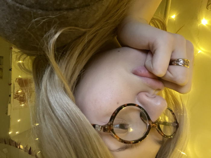 An upside-down selfie shows a person with blonde hair, wearing tortoiseshell glasses. The person's hand is raised, with a ring visible on one finger. The hand appears to be near the mouth, possibly covering it or touching the lips. Behind the person, there's a wall with a light-colored wall hanging. Strings of fairy lights decorate the background, creating a soft, warm glow. The overall image has a slightly distorted perspective due to the upside-down orientation.