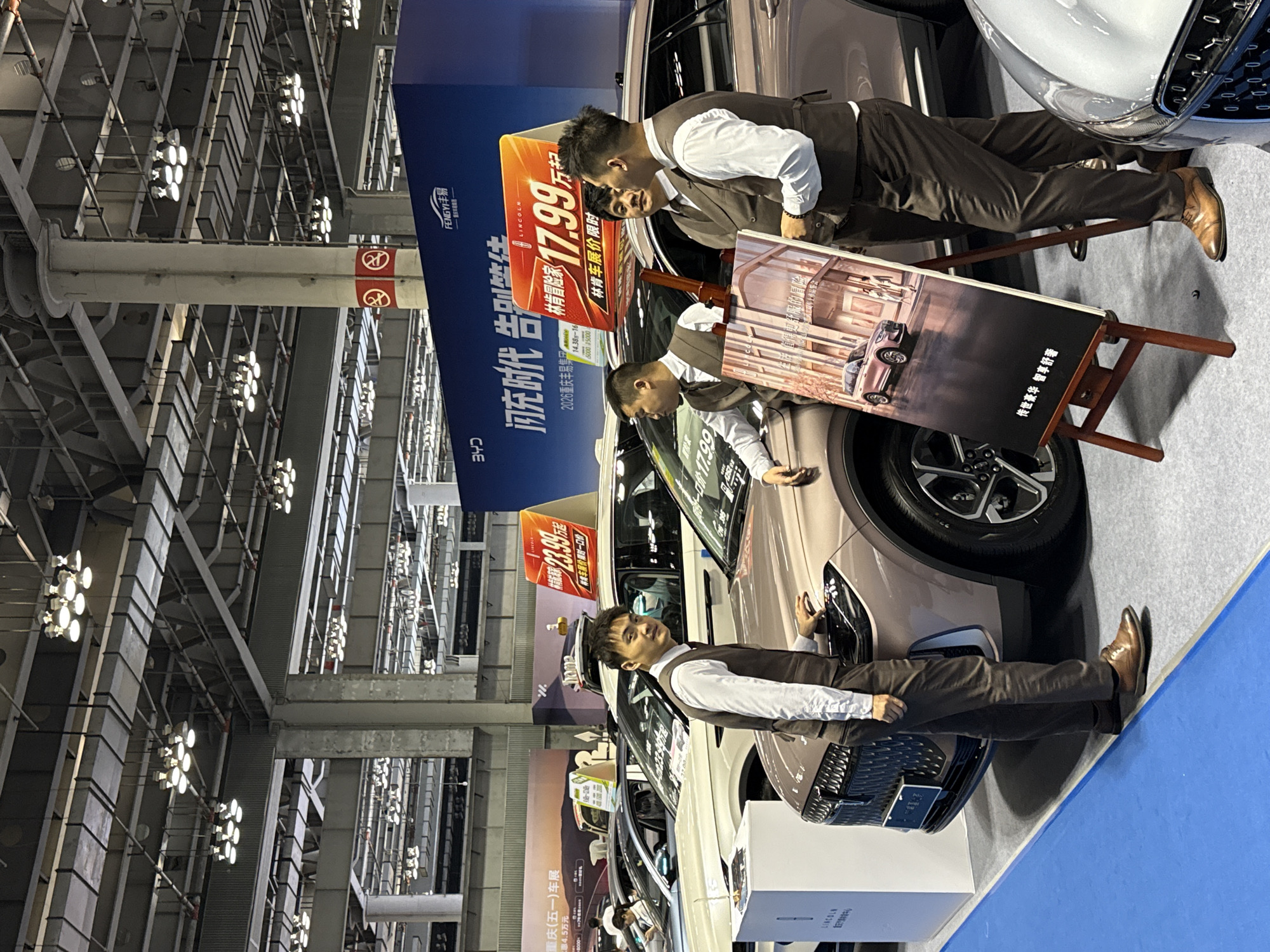 This image depicts an indoor car exhibition. The setting appears to be a large exhibition hall with a high ceiling featuring a network of metal structures and lighting fixtures. Three men are visible, all dressed in brown suits with white shirts, positioned around a vehicle. The car is light brown in color, and there is a promotional stand beside it with the name "Lincoln" displayed. The stand advertises prices and promotions such as "17.99" and "23.99". One man is standing near the front of the car, possibly engaging with the vehicle. The other two are near a sign displayed on an easel, apparently discussing or pointing towards it. The sign features an image of the same car. The overall lighting is bright, emphasizing the cars and displays. The floor is covered in a blue carpet. The image contains text in Chinese characters, with the word "Lincoln" visible in English. The location is tagged as Chongqing (五一)车展, which translates to "Chongqing (May 1st) Auto Show".