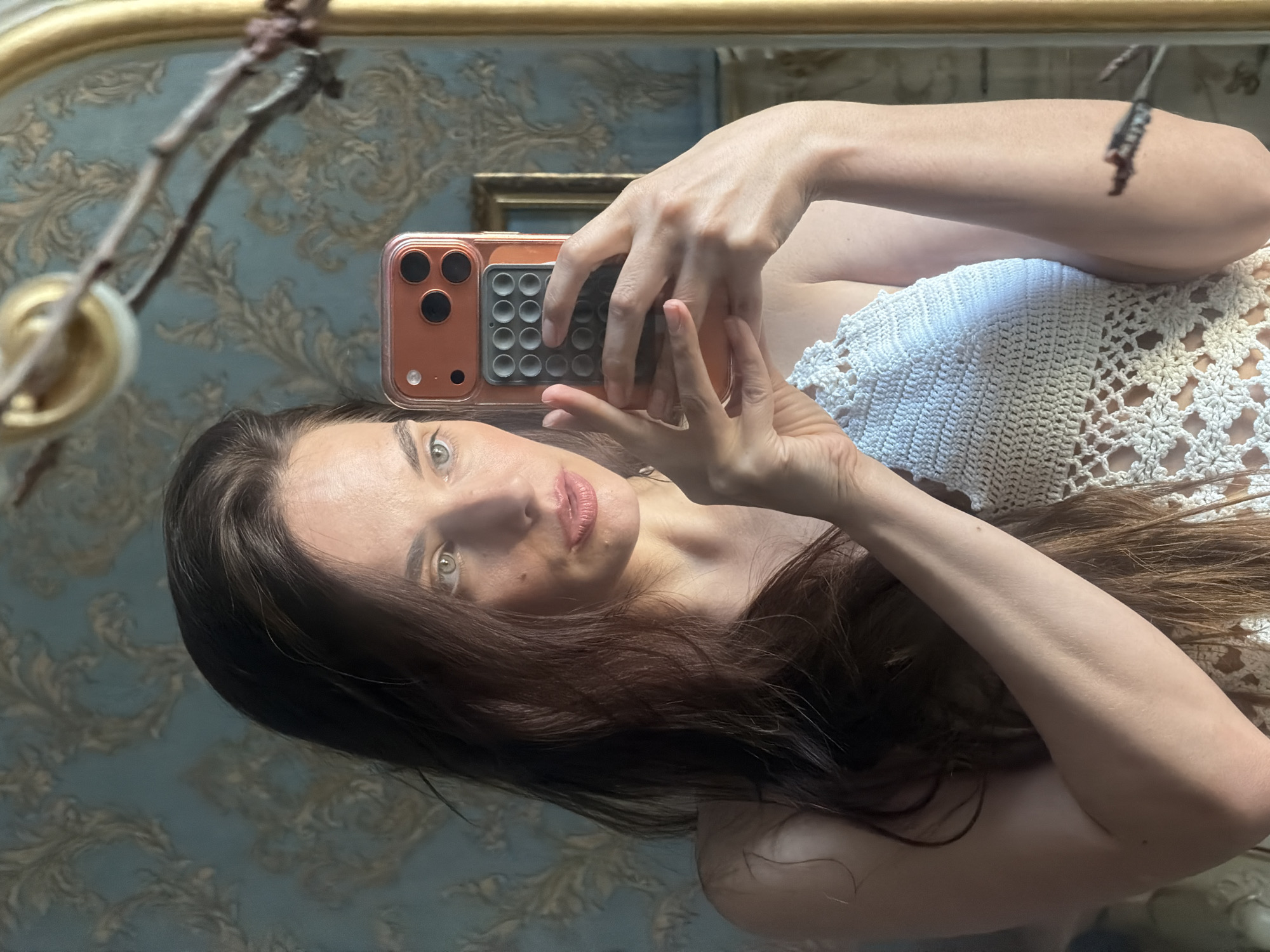 The media file is a vertical, slightly blurred selfie taken in a mirror. The subject is a young woman with long, dark brown hair and light eyes. She is wearing a white, crocheted top. Her right arm is raised, holding a cell phone, which is angled to capture her reflection. The phone is in a bright orange case with a textured grid on the back.
The background is a wall with ornate, light blue and gold patterned wallpaper, suggesting an indoor setting with a decorative aesthetic. A gilded mirror frame is partially visible at the top of the image, and a branch with some dried leaves hangs from the left side, possibly part of a chandelier or wall decoration. The lighting appears to be natural, casting soft shadows and highlighting the texture of the wallpaper and the woman's skin. The overall mood is casual and self-reflective. There is no visible text in the image. The location context provided is Ufa, Russia, but there are no specific visual cues within the image to confirm this.