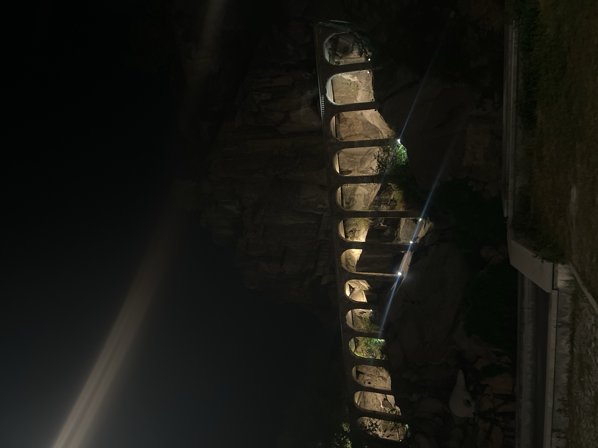 The image shows the Roman aqueduct in Plovdiv, Bulgaria, at night. The aqueduct is made of stone and consists of a series of arches illuminated by lights. It is built into a rocky hill. To the right of the aqueduct, there is some greenery and what looks like a retaining wall or structure. The left of the image is predominantly dark, indicating nighttime. There is a lens flare originating from the left side of the image.