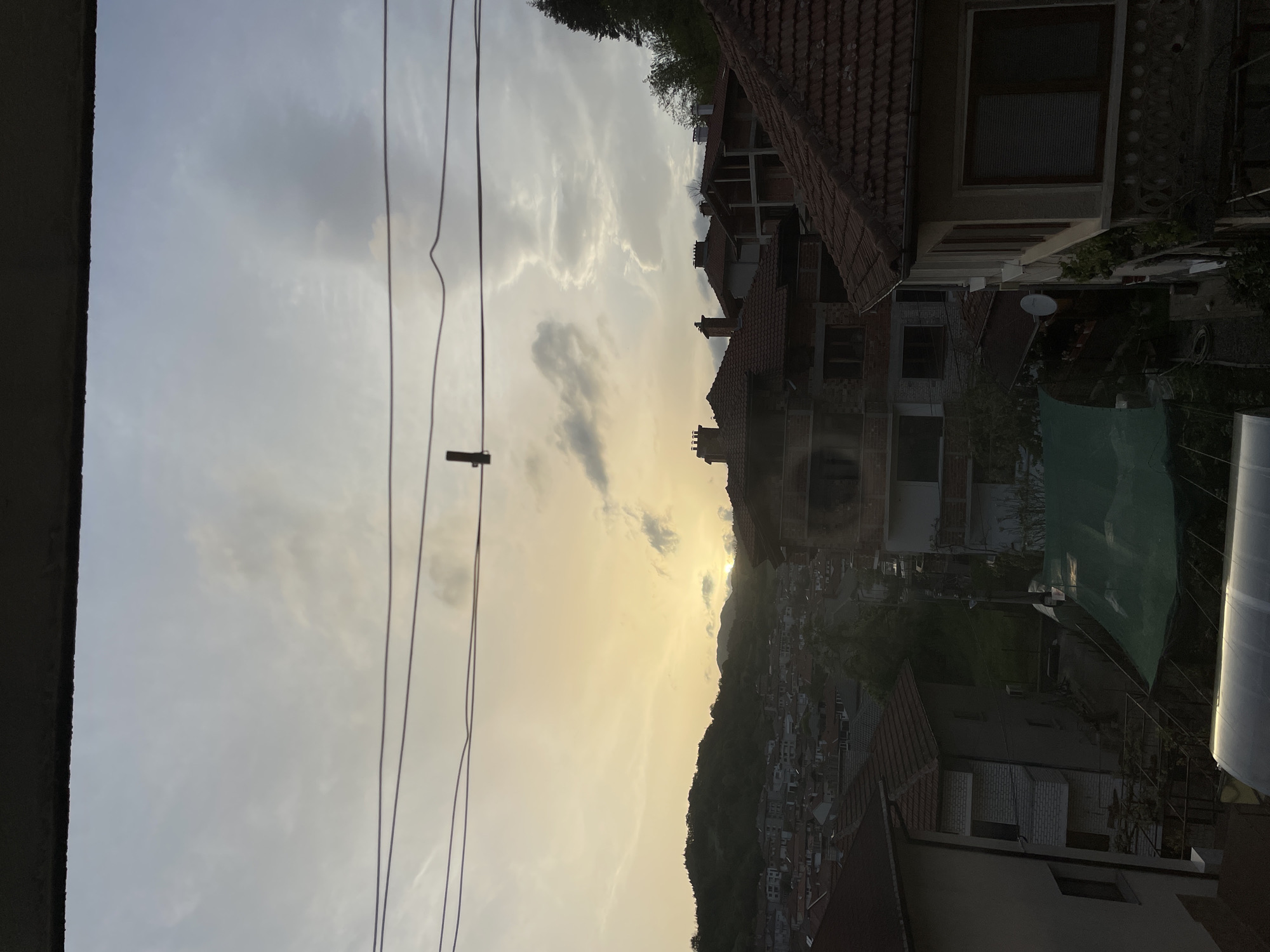The image shows a view overlooking Zlatograd, Bulgaria, at sunset. A silhouetted mountain ridge is visible in the distance with the setting sun breaking through clouds above it. Foreground objects include power lines and the rooftops of several multi-story buildings, some under construction. There is evidence of a garden area with green mesh. The sky is overcast with thin clouds.