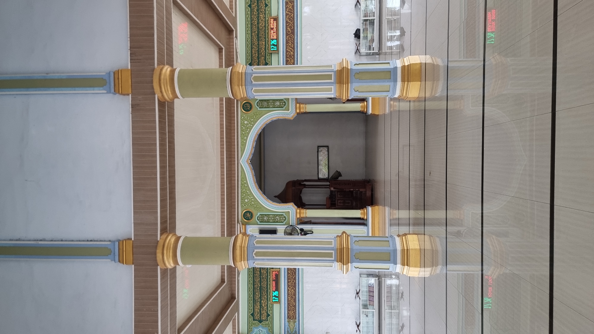 The image shows the interior of a mosque, possibly in Meunasahlhok, Indonesia, with a focus on architectural elements. Two large, decorative pillars with gold, green, and blue accents stand prominently in the foreground. Behind them, an arched entryway leads to a darker interior space where a wooden pulpit, known as a mimbar, is visible. The floor is tiled with a reflective surface that mirrors the pillars and the arched entrance. Digital displays showing prayer times are mounted on the walls on either side of the entryway, with Arabic calligraphy above them. The overall environment appears clean and well-maintained, with white tiled walls and a beige, textured ceiling. There are no people visible in the image.