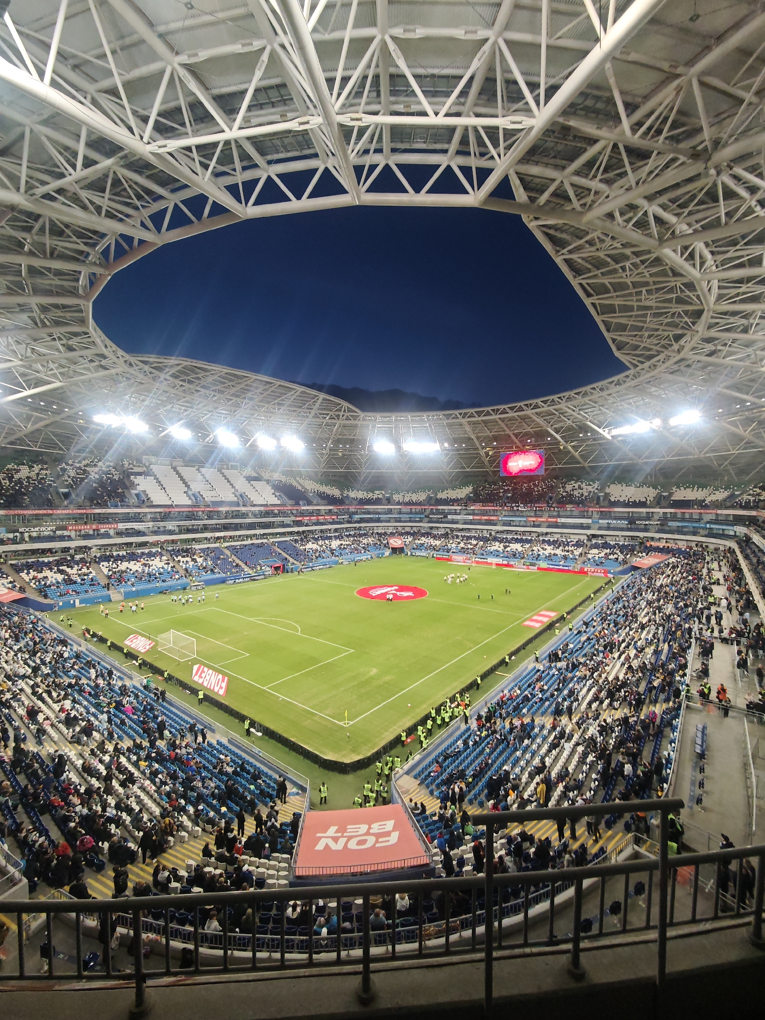 The image shows a soccer match at night inside a stadium in Novokuybyshevsk, Russia. The stadium is packed with spectators in tiered seating around a green soccer field. Players in white and blue uniforms are visible on the field. Above the stadium, there is a complex white steel structure creating an open roof with a view of the dark night sky. A large video screen is visible in the distance. Several bright lights illuminate the field. A banner with the word "FONBET" is visible near the bottom of the image. A metal railing is present in the foreground.