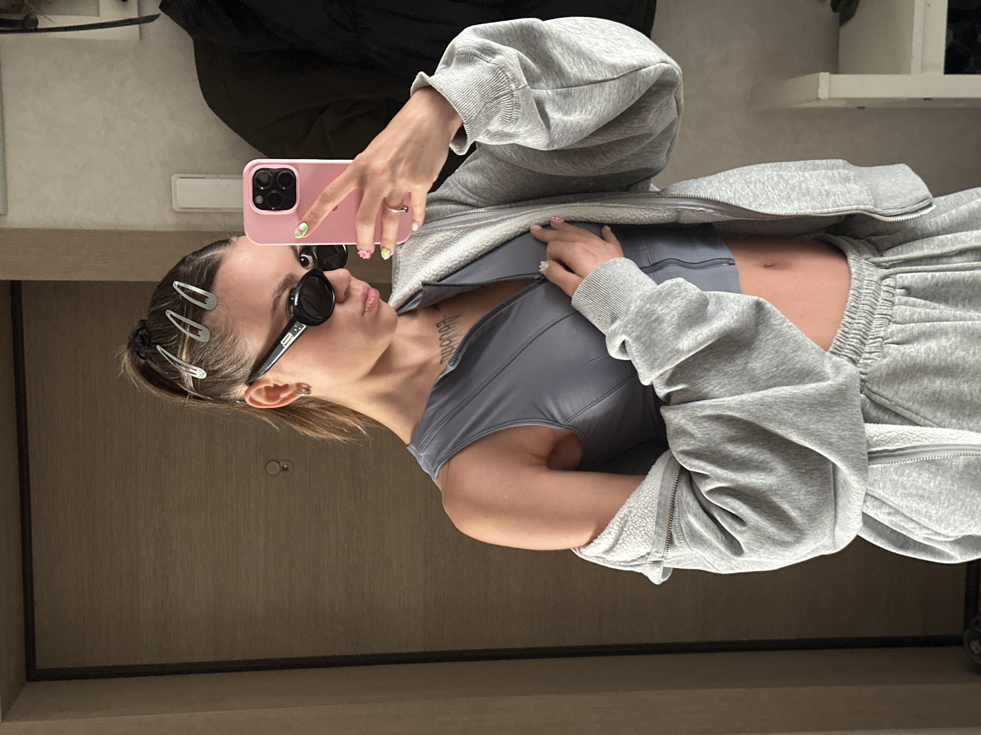 A young woman is taking a mirror selfie in what appears to be an apartment hallway. She is wearing a grey athleisure set consisting of a zip-up hoodie over a cropped tank top and sweatpants. She has black sunglasses on and her hair is pulled back in a ponytail with silver bobby pins. Her right hand is holding a pink smartphone, and her left hand is resting on her chest. The background shows a light brown door and a white shelf unit. The image is set in Moscow, Russia.