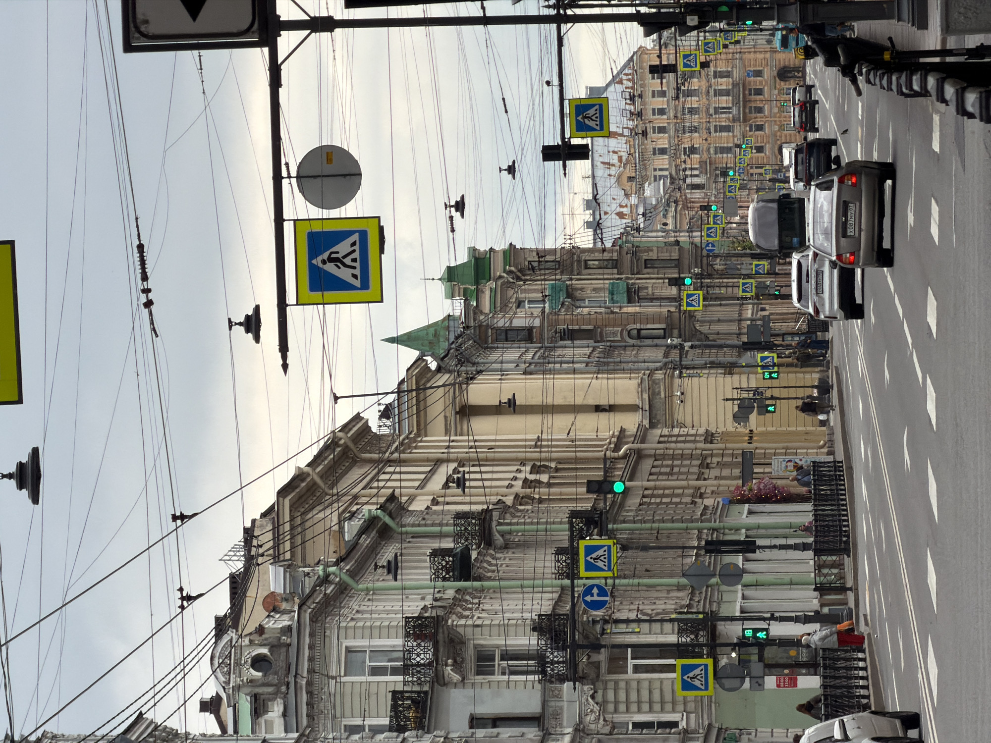 The image captures a street scene in Saint Petersburg, Russia, notable for its mix of old architecture and modern infrastructure. The street is lined with buildings exhibiting ornate facades, some with visible wear and tear, characteristic of older urban structures. The architectural style includes details such as balconies, decorative moldings, and varying rooflines. Overhead, a dense network of wires spans the street, typical of urban environments with extensive public transportation and utility systems. Numerous street signs, including pedestrian crossing symbols, are mounted on poles along the street. Traffic flows along the road, with several cars visible, some in motion. Sidewalks are present, though details about pedestrian activity are limited. The overall lighting suggests a daytime scene under overcast conditions.