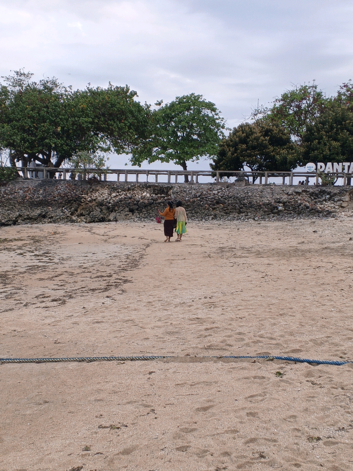 The image depicts a beach scene in Denpasar, Indonesia, with two individuals walking away from the camera on the sand. The sky is overcast. In the background, there is a wall constructed from rocks and a wooden railing. Behind the railing, trees with green foliage are visible. To the right, a sign that reads "PANTAI" (which means "beach" in Indonesian) with the Aqua logo is partially visible. A thick blue rope lies horizontally across the foreground of the sandy beach.