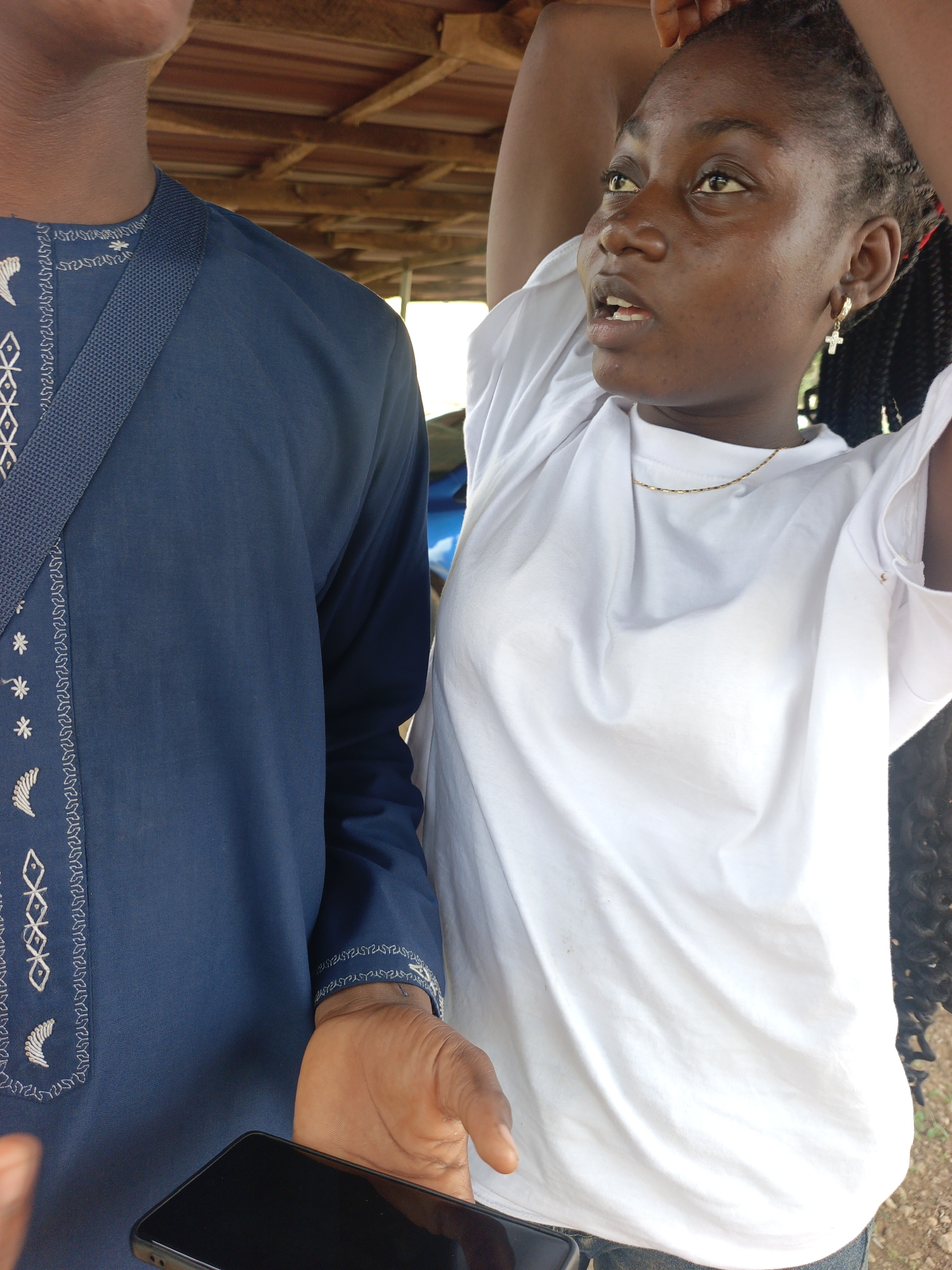 In an outdoor, under-shelter setting, a person in a navy blue tunic with white embroidered patterns is partially visible on the left side of the frame. Their right hand, holding a black smartphone, is extended towards the bottom of the image. Standing to their right is a young woman with dark skin and braided hair, wearing a white t-shirt and a thin gold necklace. She has a cross-shaped earring in her right ear and her hands are raised above her head. Her mouth is slightly open as she looks upwards and to the right. The background shows a wooden ceiling structure and some greenery.