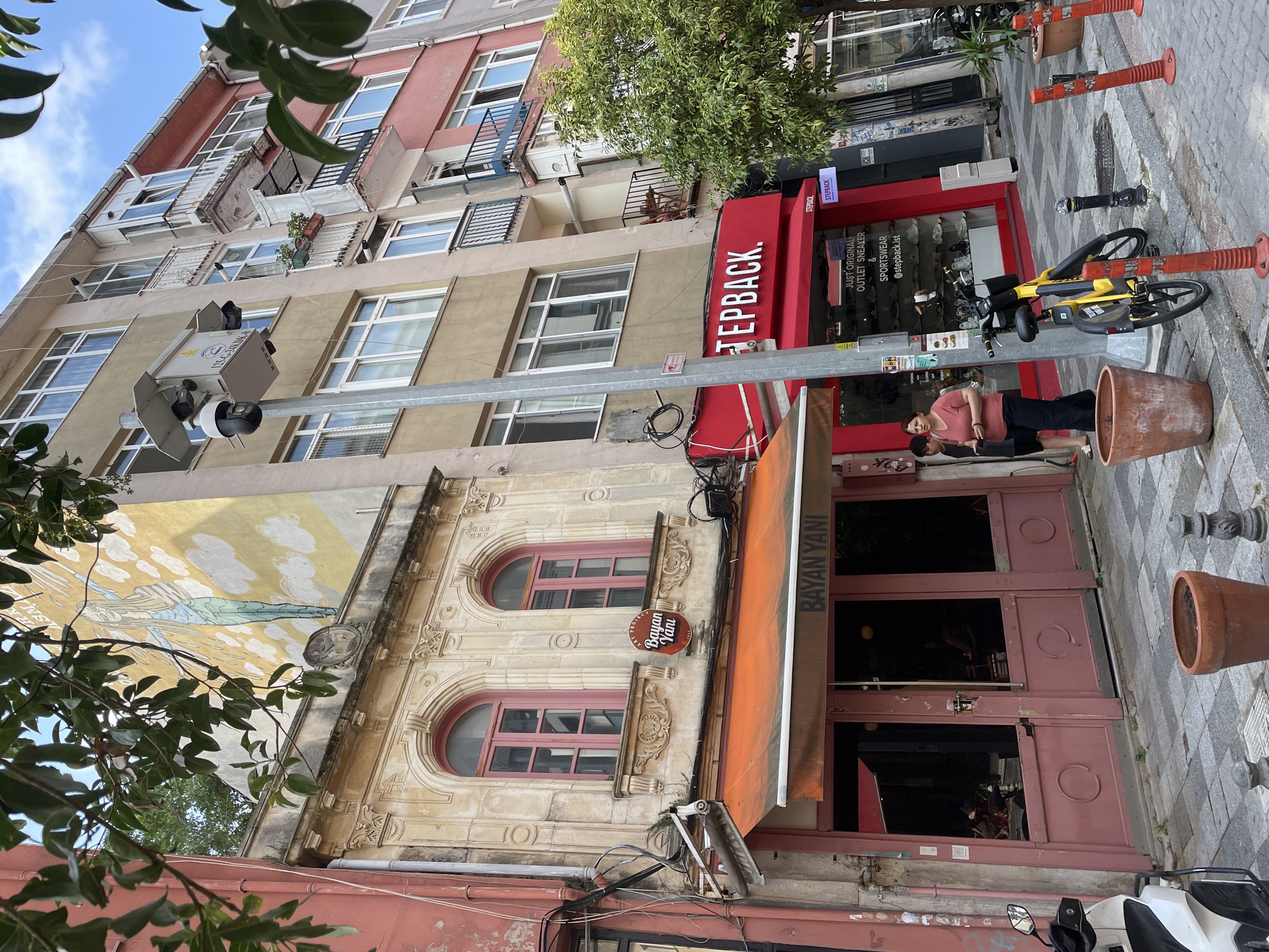 The image captures a street scene in Kadıköy, Turkey, on a bright, overcast day. In the foreground, a white scooter is parked on the left. A young woman in a pink shirt and black pants stands in front of a shop with an orange awning. Beside her, a yellow bicycle is parked vertically, leaning against a pole. The pole itself is a traffic light pole with several security cameras attached. The street is paved with cobblestones.
The shops visible are "BAYAN YANI" with an orange awning, and a red storefront labeled "STEPBACK." The "STEPBACK" store appears to be a retail establishment selling sneakers and sportswear, with merchandise visible through its windows. Further down the street, other shopfronts and residential buildings can be seen. A large tree with green foliage grows on the right side of the frame. The buildings are multi-story, with windows and balconies. A mural of a winged figure is painted on the side of one of the buildings. Traffic cones are placed along the sidewalk.