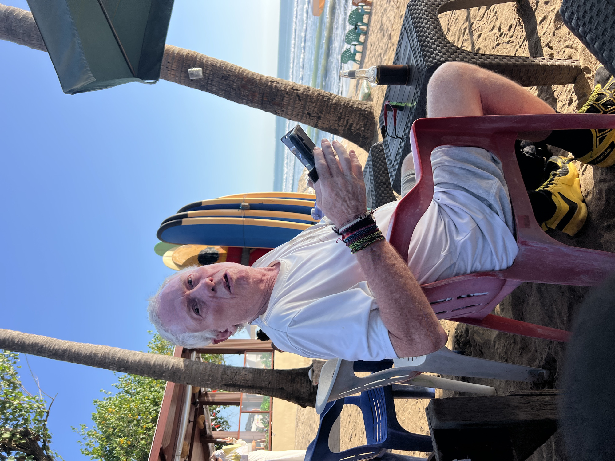 A man with white hair and fair skin is sitting in a red plastic chair on a sandy beach. He is wearing a white t-shirt and light-colored shorts. He is holding a smartphone in his right hand and appears to be looking at it. His left arm is resting on the armrest of the chair and has several braided bracelets on his wrist. On a woven plastic table in front of him, there is a pair of sunglasses, a bottle with a dark liquid in a brown holder, and what appears to be a cup. Behind him, a palm tree trunk is visible, with several surfboards leaning against it. Further in the background, the ocean with waves can be seen, along with several green beach chairs and an umbrella. The man is looking slightly off to his left with a neutral expression. The lighting suggests it is daytime, likely late afternoon given the shadows. The location is identified as Kuta, Indonesia.