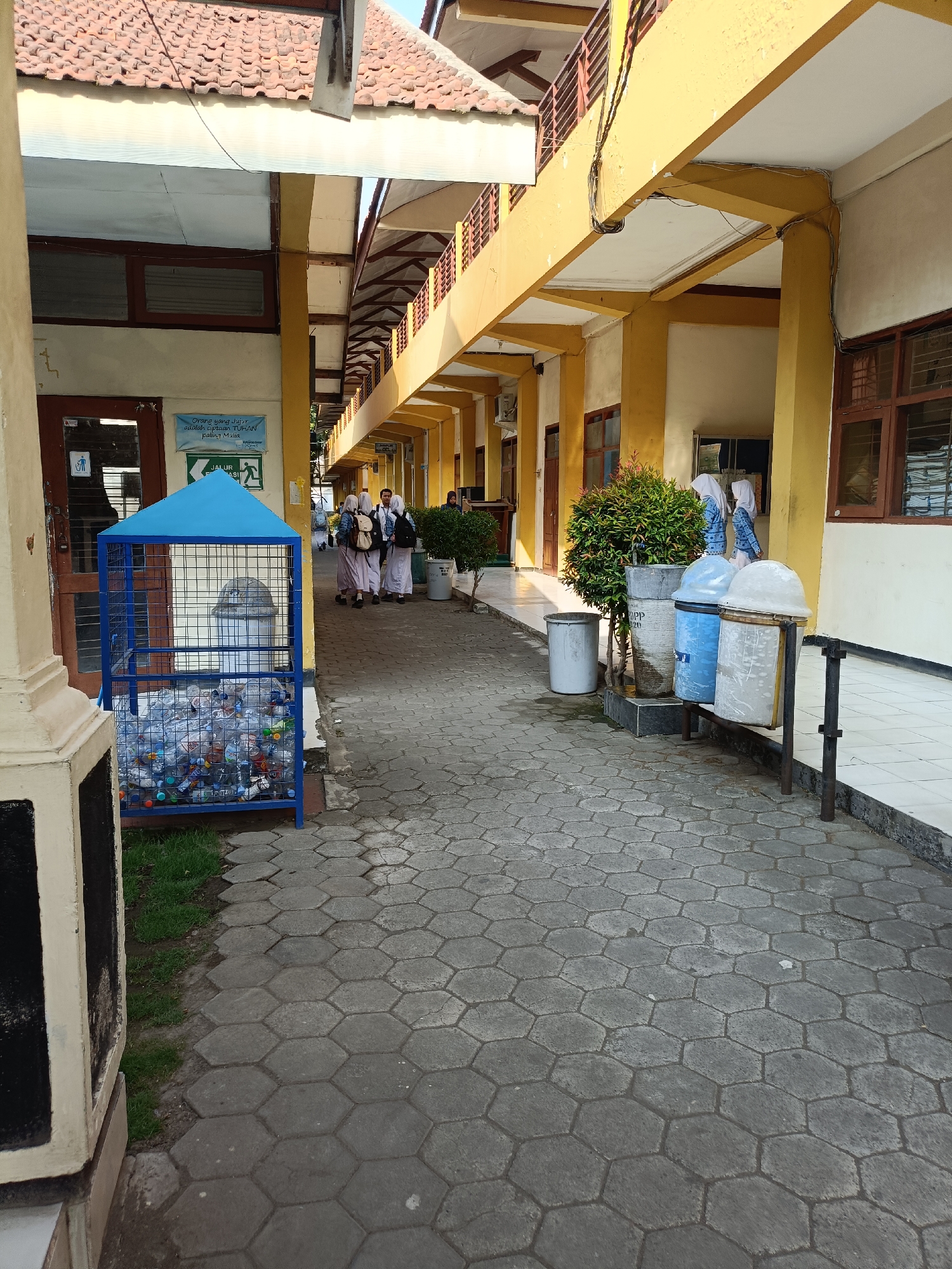The image depicts a school hallway with a covered walkway. The walkway is paved with hexagonal gray tiles. On the left side, there is a blue metal recycling bin filled with plastic bottles. Next to it, a brown wooden door with glass panels is visible, and above it, a sign with text and an icon. Further down the hallway, three female students wearing hijabs and uniforms are walking away from the camera. They are carrying backpacks. On the right side of the walkway, there are several trash cans. The hallway is lined with yellow pillars supporting the roof. The environment appears to be an outdoor school corridor in Kediri City, Indonesia, under a bright sky, suggesting it is daytime.