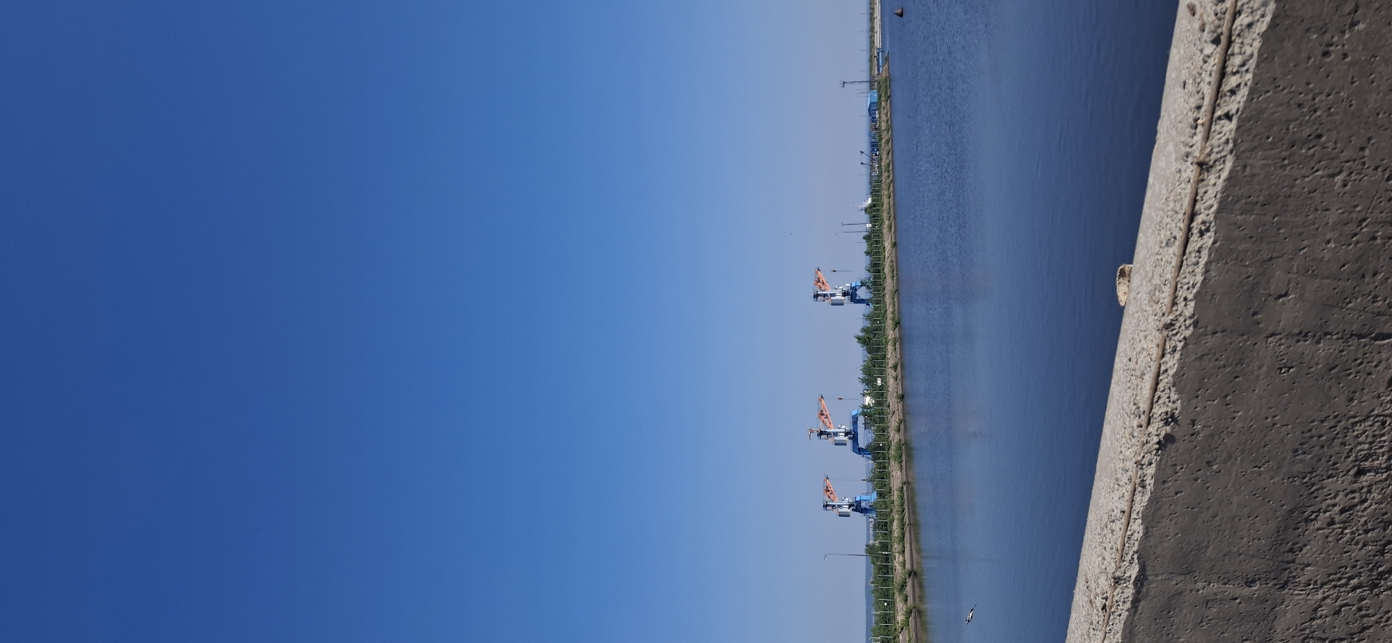 The image shows a waterfront scene in Chaikovsky, Russia. Three blue and orange cranes are positioned along the water's edge. The sky is clear and blue. A concrete wall is in the foreground. The water is calm. A thin strip of green vegetation runs along the waterfront. There are structures in the distance.
