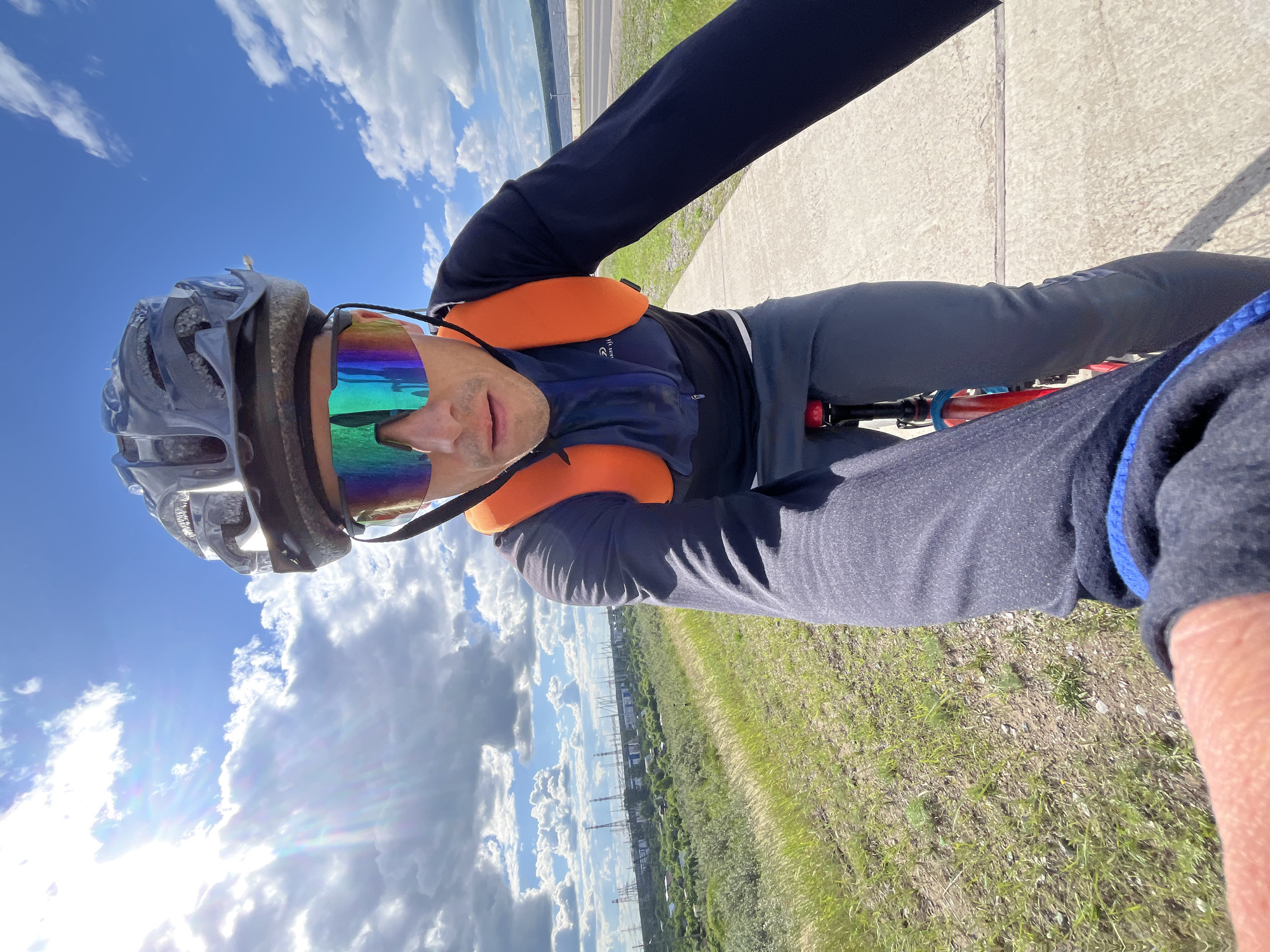 A man wearing a cycling helmet and reflective sunglasses rides a bicycle on a paved path. He is dressed in a dark blue long-sleeved shirt, grey pants, and an orange vest. The man is looking forward, as if at the camera. The background shows a grassy slope leading down to a landscape with a body of water, a road, and industrial structures. The sky is blue with scattered clouds and the sun is shining brightly. The scene is likely taking place in the city of Chaikovsky, Russia, given the industrial infrastructure visible in the background which resembles the Kamskaya HPP.