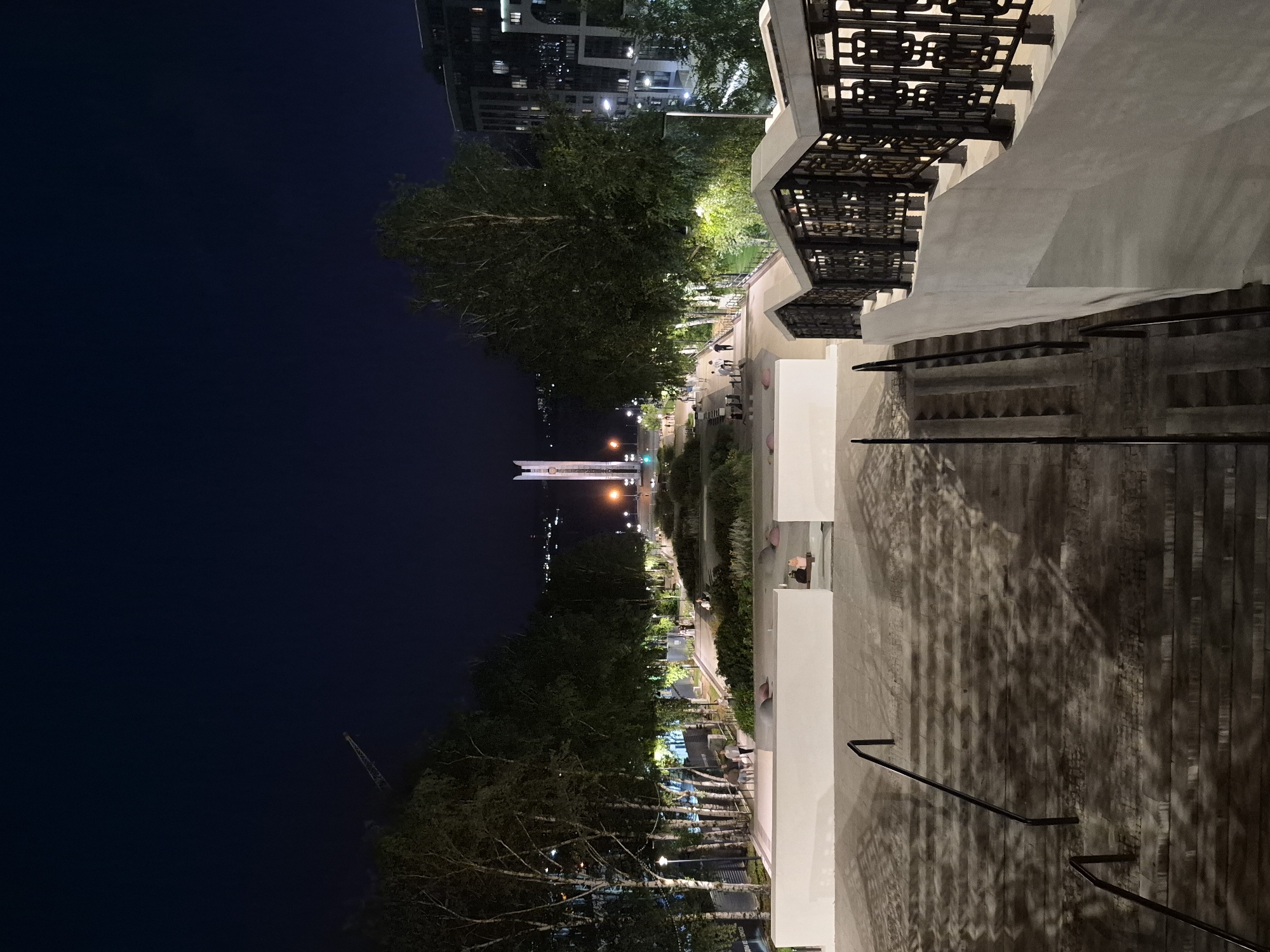 The image depicts a night scene of the Monument of Friendship of the Peoples in Izhevsk, Russia. A wide, illuminated stairway with ornate black railings leads down from the foreground. The stairway casts long shadows, suggesting artificial lighting. At the bottom of the stairway, a park-like area is visible with trees and benches. Several people are scattered throughout this area, some sitting and others walking. Further in the distance, a tall, slender monument with a pointed top stands illuminated against the dark night sky. Trees line the pathways, and streetlights provide further illumination to the scene. Modern buildings with lit windows are visible on the right side of the image. The sky is a deep, dark blue, indicating it is nighttime. A construction crane is faintly visible in the upper left corner against the sky.