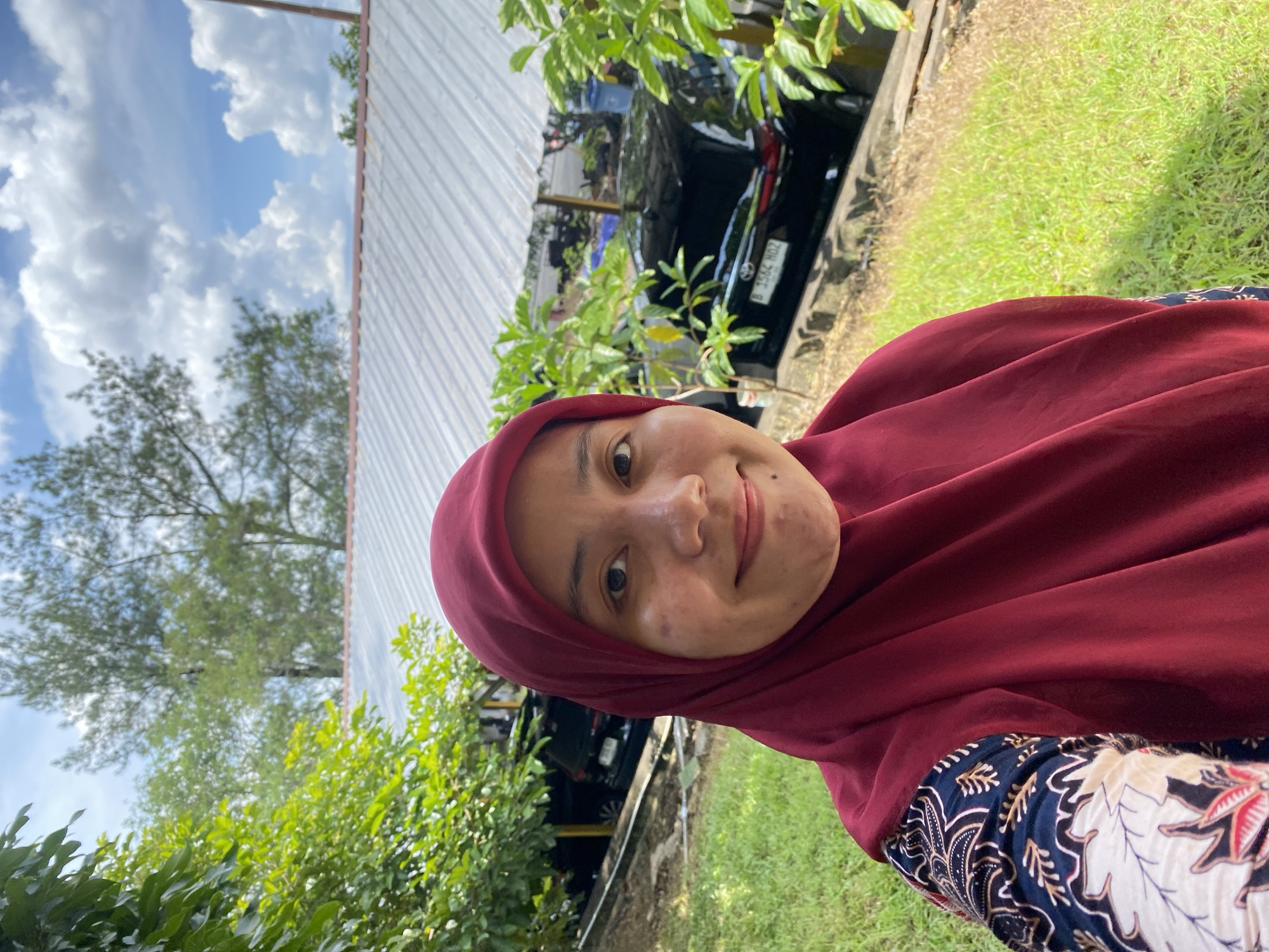 A woman wearing a maroon hijab and a patterned shirt takes a selfie outdoors. She is smiling and looking at the camera. In the background, there is a building with a corrugated metal roof, lush green trees, and a parked black car. The scene appears to be in an outdoor setting, possibly a parking area or a public space in Banjarbaru, Indonesia. The lighting suggests it is daytime, with sunlight filtering through the clouds.