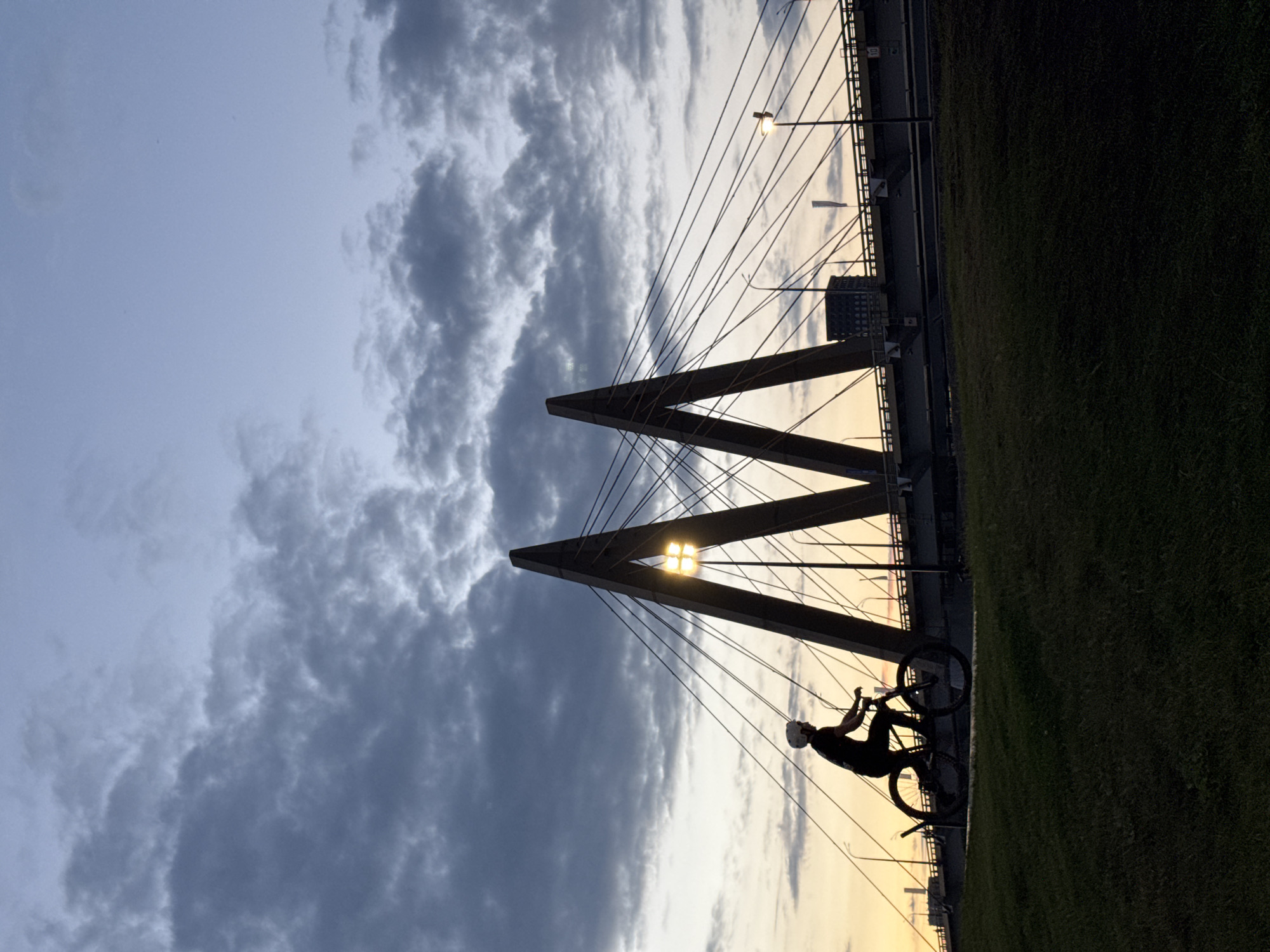 A silhouette of a cyclist wearing a helmet rides a bicycle in front of a cable-stayed bridge at dusk. The sky is a gradient of blue and yellow, with dark clouds visible in the upper portion. The bridge features two large, triangular pylons that support the cables. Streetlights are visible along the bridge, and a tall building can be seen in the background. The foreground consists of a grassy embankment. The image appears to be taken from a low angle, emphasizing the scale of the bridge and the cyclist. This is the Millenium Bridge in Kazan, Russia.