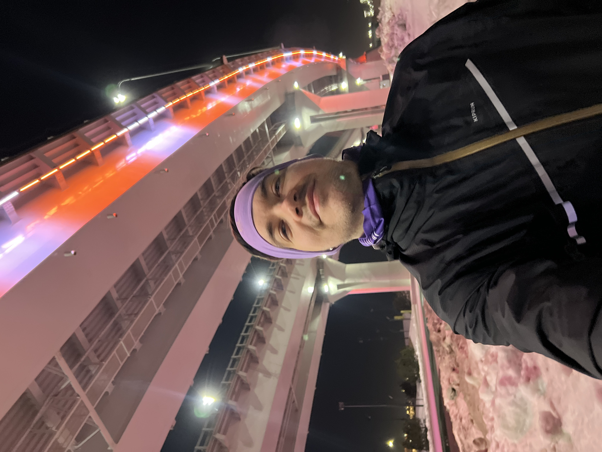 The image depicts an adult male taking a nighttime selfie in a snowy, urban environment. The male, appearing to be in his late 20s to early 30s with light skin and stubble, is looking directly at the camera with a subtle smile. He is wearing a purple athletic headband, a purple neck gaiter, and a black jacket with a gold zipper and a "KIPRUN" logo visible on the chest.
Behind him, a massive, multi-tiered concrete bridge or overpass structure dominates the background. The bridge is extensively illuminated with integrated LED lighting, displaying vibrant orange and purple/pink hues along its visible side, while white lights illuminate its underside. Multiple support pillars and a roadway with railings are discernible within the structure. Several streetlights with bright white illumination are visible under and beyond the bridge, contributing to the scene's lighting. The ground below is covered in significant accumulations of snow, forming irregular mounds, some of which reflect the colorful light from the bridge. Distant city lights and sparse vegetation are visible on the horizon.
This scene is located near the Millyenium Bridge (Millennium Bridge) in Kazan, Russia, identifiable by its distinctive multi-level architecture and prominent colorful nighttime illumination.