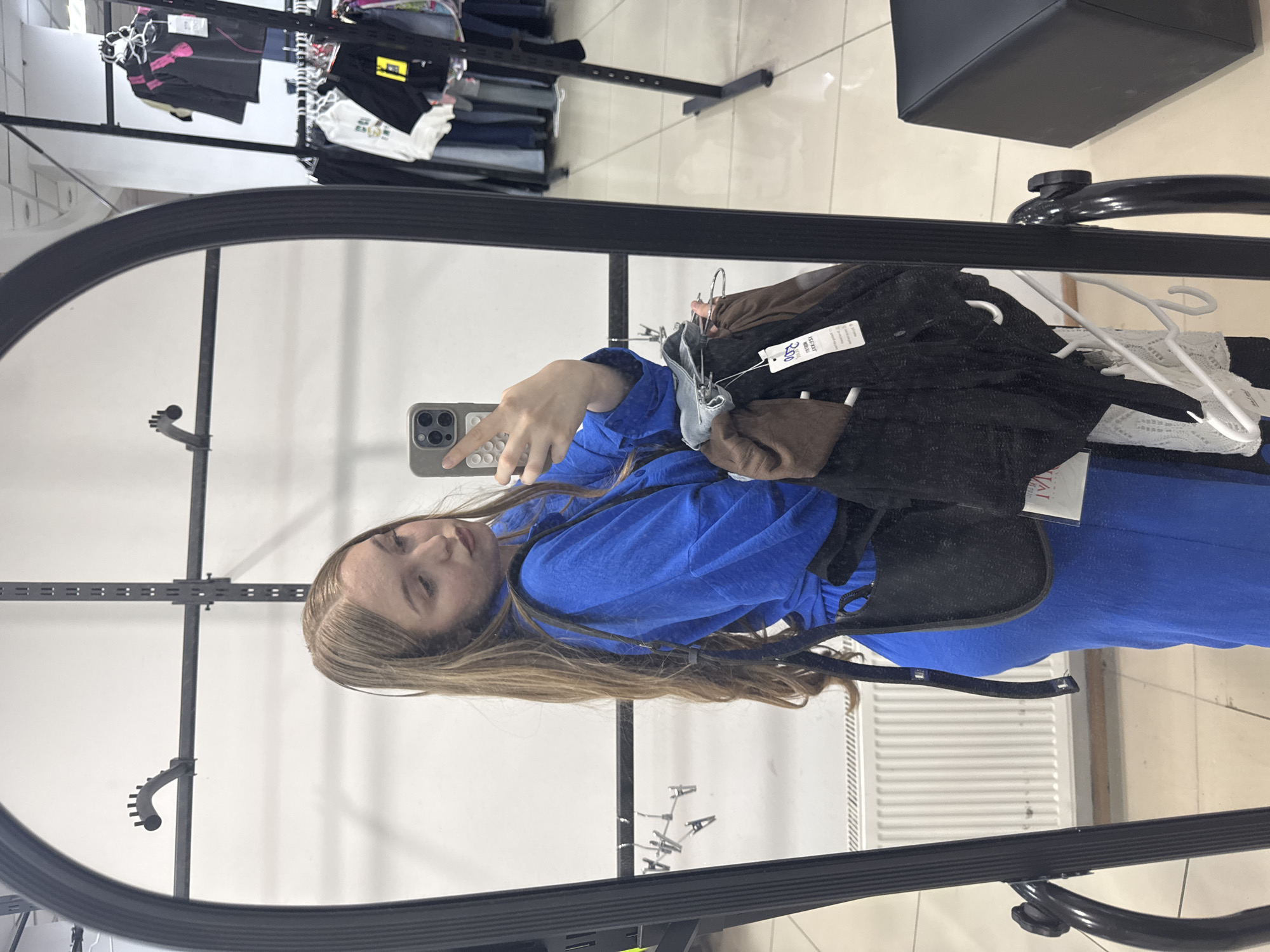 A young woman with long blonde hair is taking a selfie in a mirror. She is wearing a bright blue top and appears to be in a clothing store. The mirror reflects racks of clothing, including black, white, and pink garments, as well as some denim. A black stool or bench is visible in the upper right corner of the reflection. A price tag with Cyrillic text is visible on a piece of clothing. The woman is holding a smartphone in her right hand. The overall impression is casual and typical of someone trying on clothes.