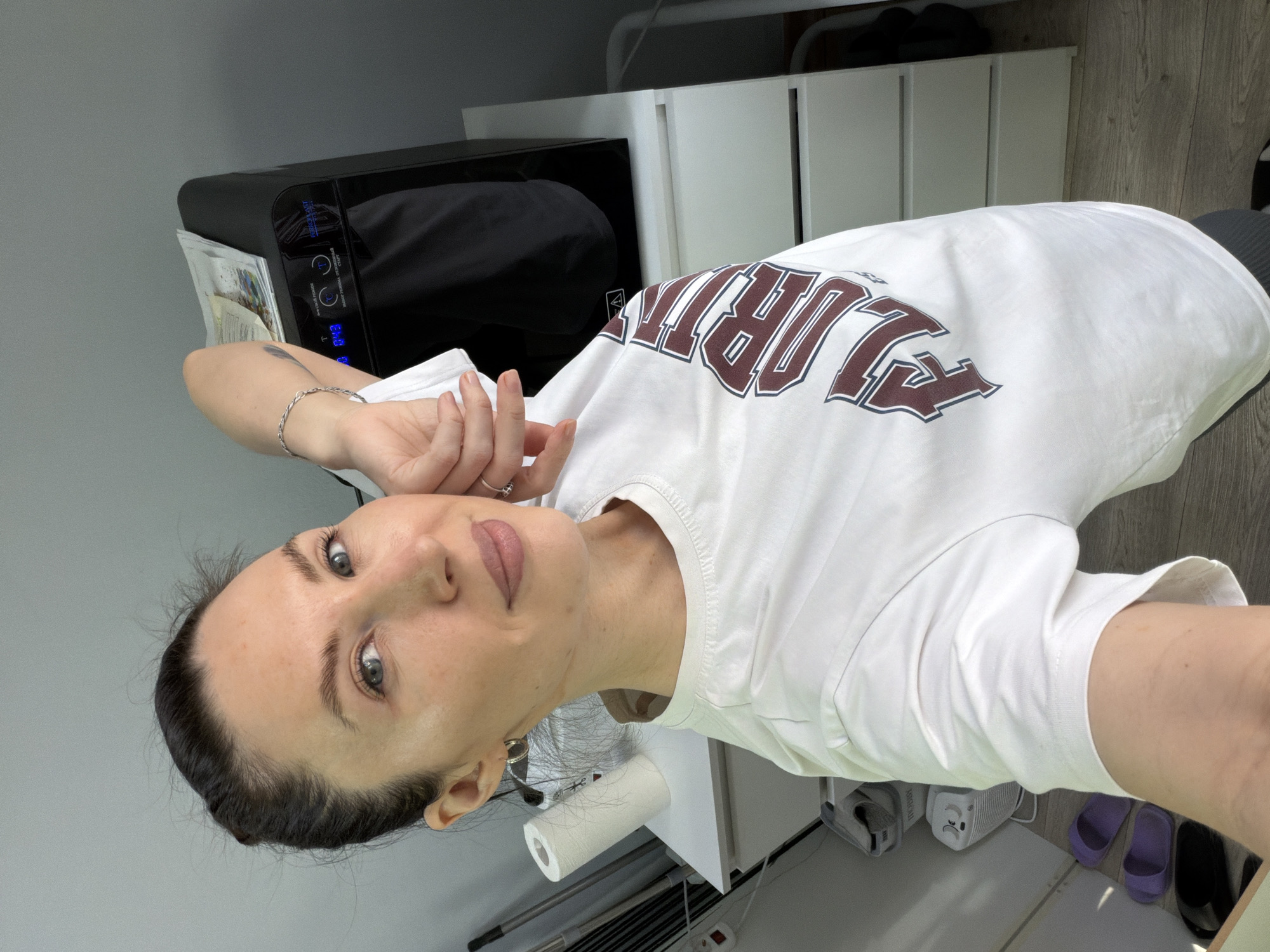 A woman with dark hair pulled back is posing for a selfie. She is wearing a white t-shirt with "FLORIDA" printed on the front in maroon lettering. She has a silver bracelet on her left wrist and a ring on her finger. The location appears to be an indoor setting, possibly a beauty salon or spa. Visible in the background are white cabinets, a black piece of equipment (possibly a sterilizer), a roll of paper towels, slippers and other miscellaneous items. The lighting is bright and appears to be artificial. This image was taken in Perm, Russia.