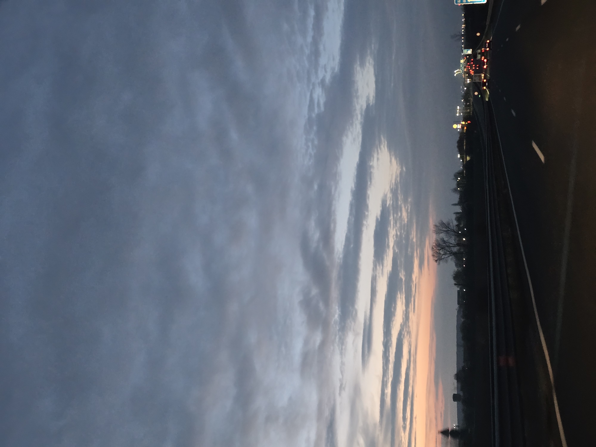 The image depicts a road at dusk or dawn, showing a highway with multiple lanes of traffic. In the distance, there are lights from buildings and vehicles, indicating an urban environment. The sky is filled with clouds, displaying a mix of gray and pale colors. The location cannot be determined, as there is no data pointing to Sofia, Bulgaria.