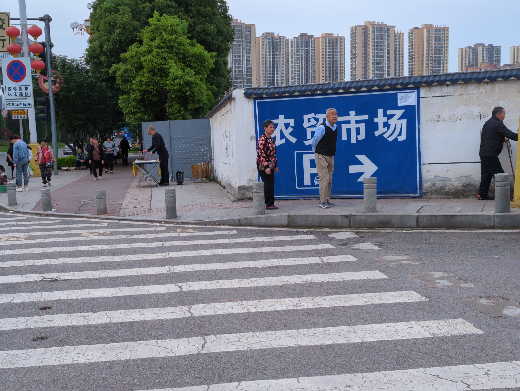 This image captures an urban street scene in Jiangjin, China, likely during the late afternoon or early evening under an overcast sky. In the foreground, a zebra crossing with worn white stripes spans the road, bordered by grey cylindrical bollards that separate the street from the sidewalk.
On the right side of the frame, a white wall features a prominent blue sign. The sign displays "农贸市场" (Farmers Market) in large white characters, accompanied by a right-pointing arrow and the letters "D" and "B." Three individuals are standing near this wall: an older woman in a floral top, an older man in a light blue shirt and dark vest, and another man in a black jacket leaning against the wall, holding a yellow stick.
Further along the sidewalk to the left, a man in black attire stands next to a small table, possibly engaging in street vending activities, with a metal trolley behind him. Several other people are walking or standing on the sidewalk, including a woman in a denim jacket. A child is partially visible on the far left.
A tall street pole on the left is decorated with multiple red lanterns and various traffic signs. These signs include a "No Parking" symbol and Chinese text such as "违停抓拍 执法路段" (Illegal parking enforced by camera, Enforcement section), "停车线内除外" (Except within parking lines), "严管路段" (Strictly managed section), and "南北大道" (North-South Avenue). Lush green trees frame the upper left of the image, behind which numerous tall, modern residential buildings are visible against the sky, indicating a densely populated area. The scene depicts everyday urban life with pedestrians and street activity.