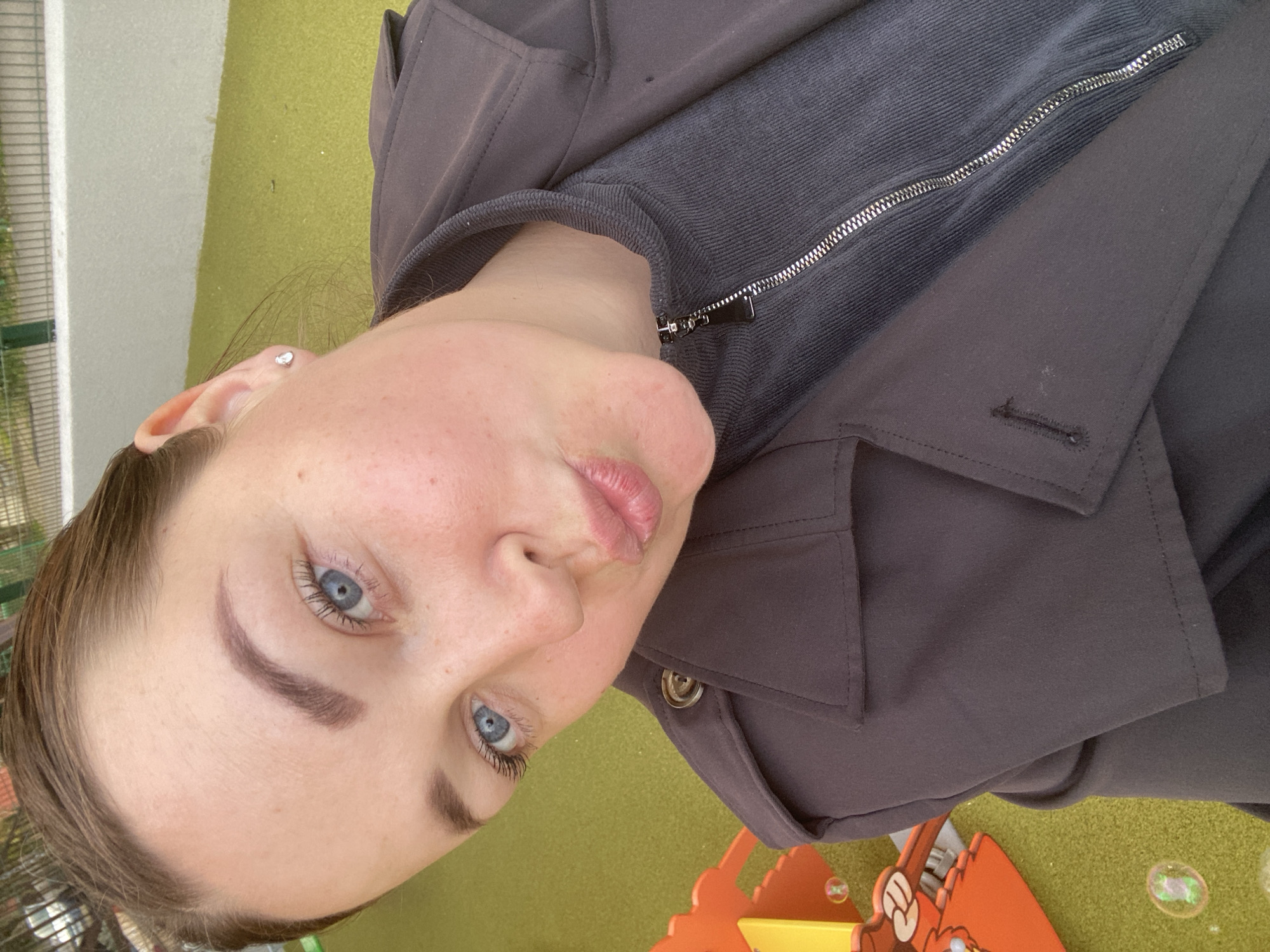 A young woman with light skin, blue eyes, and light brown hair is posing for a selfie-style photograph outdoors. She is wearing a dark gray zip-up jacket with a collared overcoat. The background appears to be a children's play area with green artificial turf, orange play equipment, and what appear to be bubbles. The location is possibly in the city of Sochi, Russia, though there is no explicit information in the image to confirm that. The woman is facing the camera with pursed lips. She has an earring in her left ear.