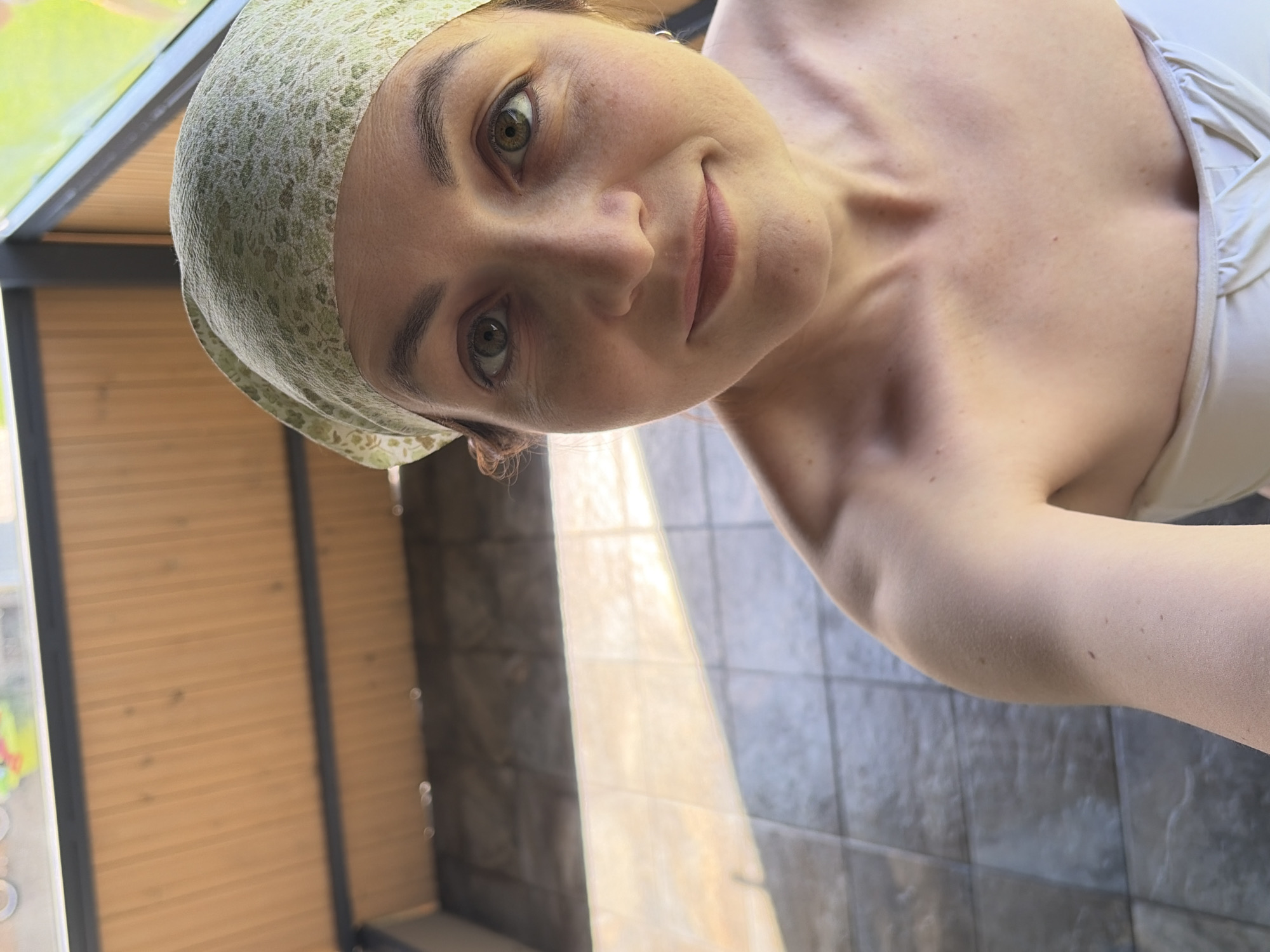 A selfie of a woman in a light-colored floral cap and a white strapless top, possibly a bathing suit. She has fair skin, light brown hair, and green eyes. She is slightly smiling. The background includes a wooden slatted wall and grey tiled wall. The location is possibly in Chaikovsky, Russia. The photo may have been taken inside of a sauna or a restroom.