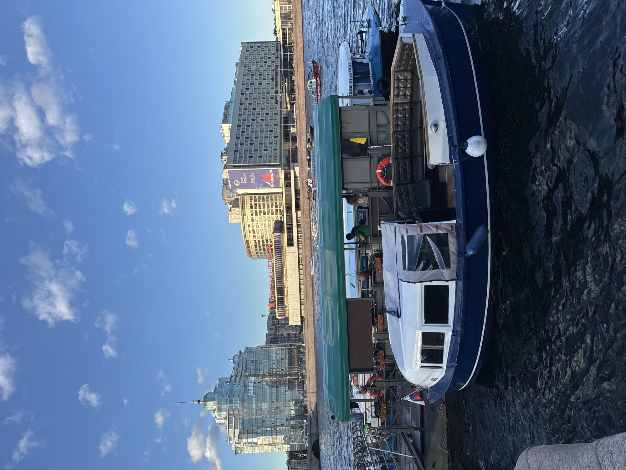 The image depicts a sunny day on a body of water, likely a river or canal, in Saint Petersburg, Russia. In the foreground, a blue and white tour boat with a green canopy is docked. The boat features seating areas with dark brown upholstered benches and a white-rimmed cabin with windows. A Russian flag is visible on the stern of the boat.
In the background, a wide river or canal separates the viewer from a cityscape dominated by modern buildings. To the left, a tall, glass-encased building with a pointed spire stands out. To the right, a larger, multi-story building with many windows is visible, featuring a prominent billboard that reads "БАНК РОССИИ" (Bank of Russia). Several smaller boats are also on the water, including a police boat with red and white markings. The sky is blue with scattered white clouds. The scene suggests a tourist area in Saint Petersburg, likely near the city center, where boat tours are a popular attraction.