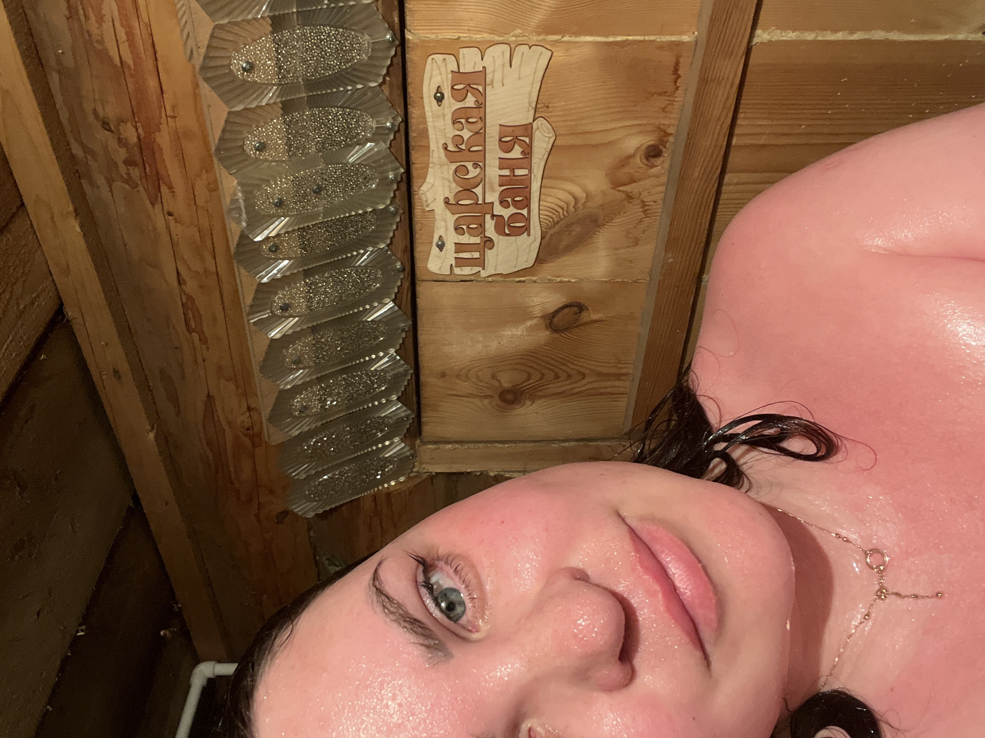 A close-up, medium shot captures a woman with dark hair, fair skin flushed red, and visible sweat droplets on her face and upper body. Her eyes, a light blue, are looking towards the camera. She is wearing a delicate gold necklace. The background consists of wooden walls and a decorative sign with Cyrillic lettering, indicating a location named "Царская баня" (Tsar's Bathhouse). The lighting is warm and appears to be from an overhead source. The overall setting suggests an interior of a sauna or bathhouse.