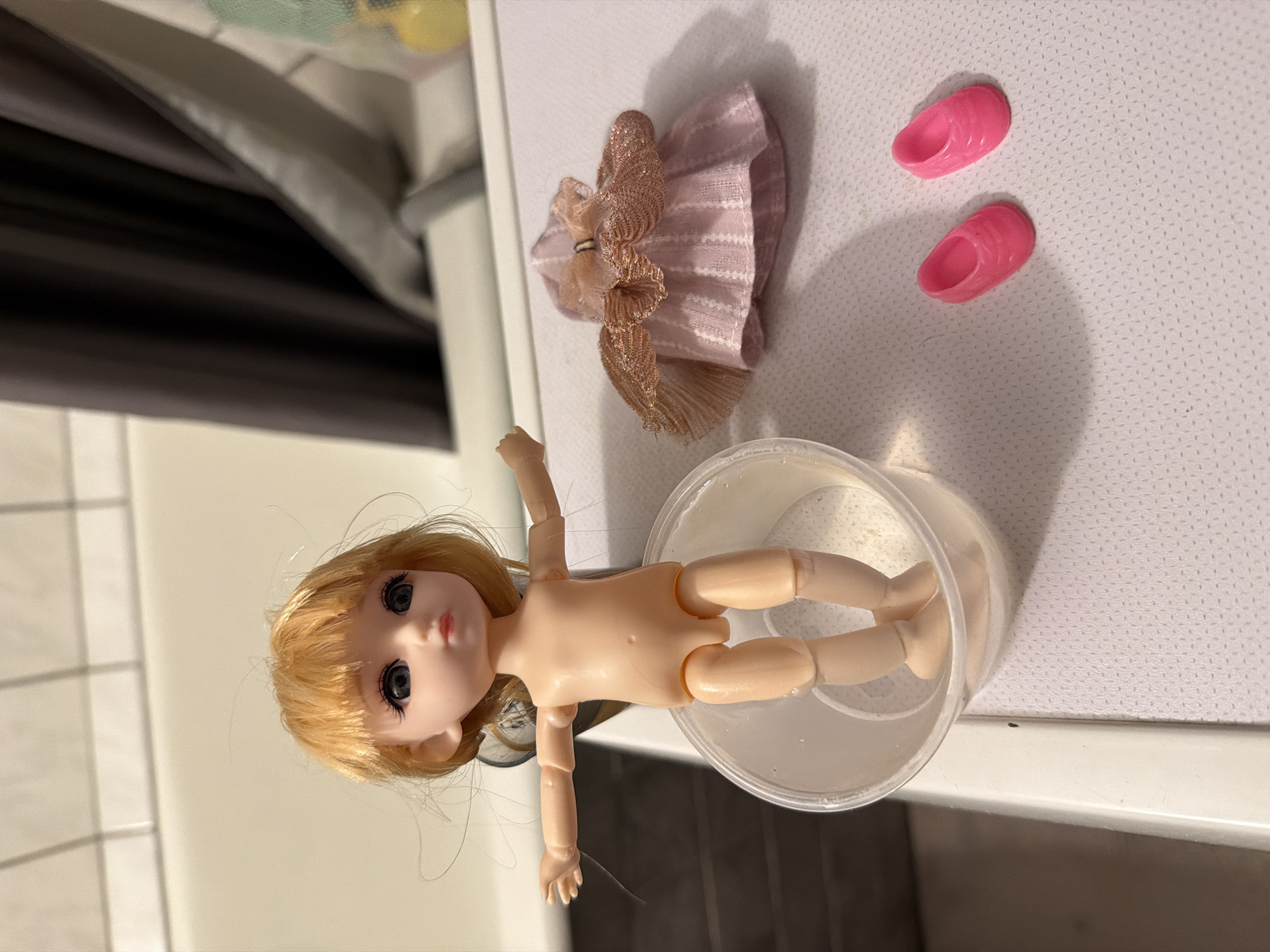 A nude doll with blonde hair lies in a clear plastic container filled with liquid. Next to the container, there is a pink dress and a pair of pink shoes. The objects are arranged on a white surface. The image was taken indoors in a room with white walls. It is not possible to determine the location as Khimki, Russia without further information.