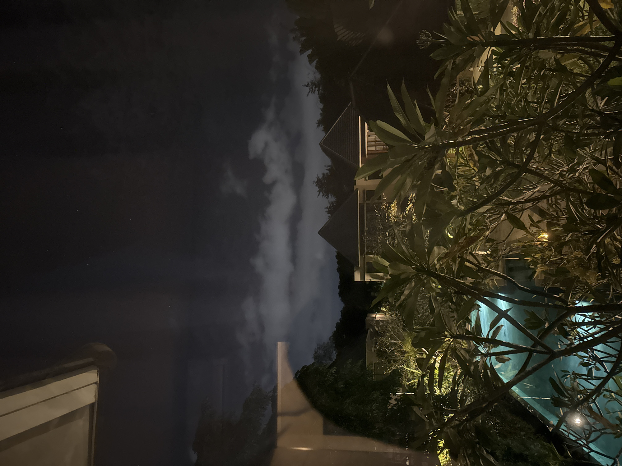 The image shows a nighttime scene in Kuta Selatan, Indonesia. A building with a peaked roof and a balcony is visible, partially obscured by trees with large leaves. The sky is dark with streaks of clouds. The lighting is dim, with artificial light illuminating the building and the leaves. A swimming pool is seen through the trees, its water reflecting a blue-green light. The photograph is taken from an elevated position, possibly from inside a building looking out towards the scene.