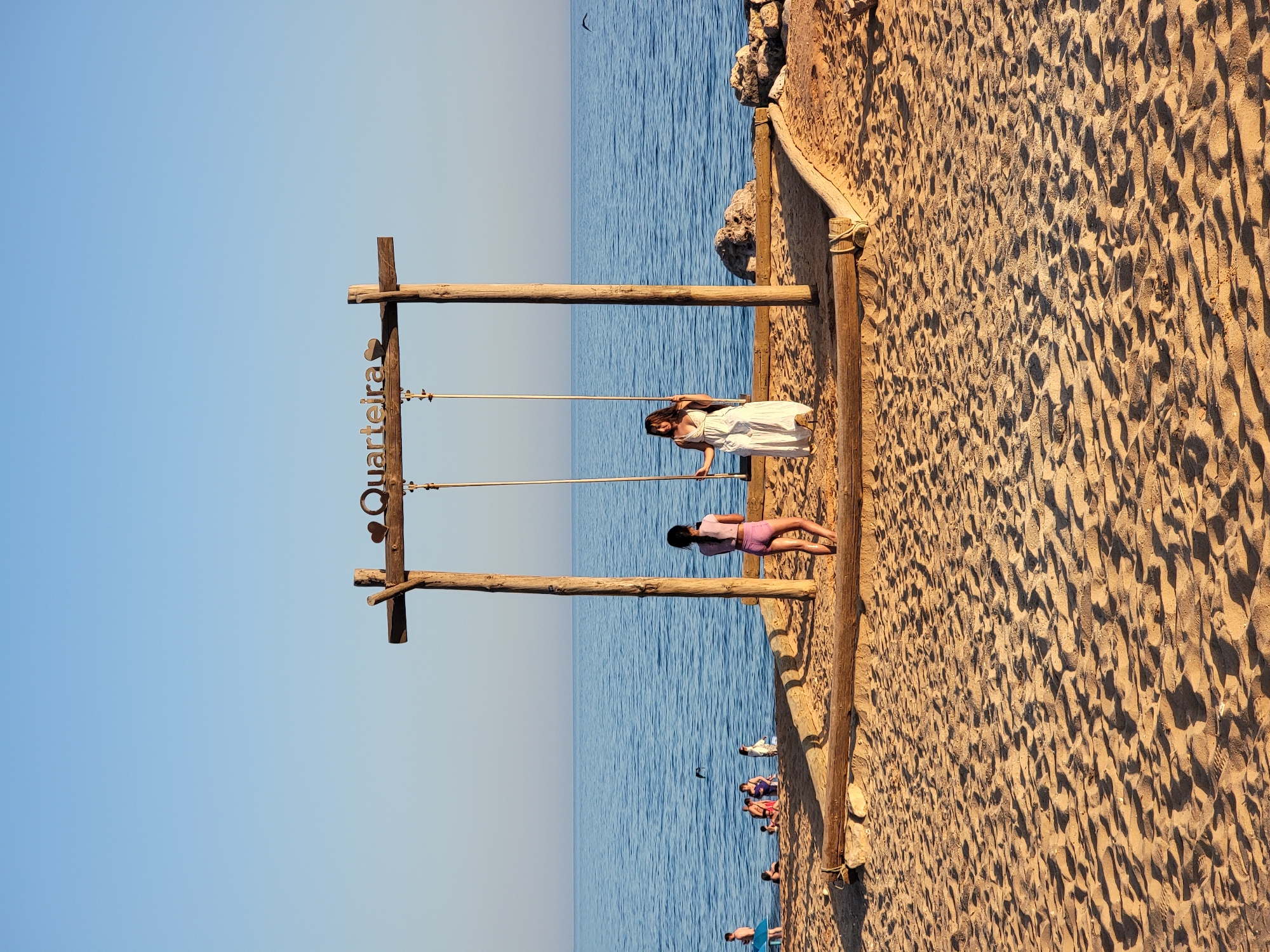 Two young women are the central subjects of this image, standing on a sandy beach in Quarteira, Portugal. The woman on the right, wearing a long white dress, is seated on a swing hanging from a rustic wooden frame. Her hair is dark and falls past her shoulders, and she is looking towards the left. The woman on the left, appearing younger, is wearing a purple two-piece swimsuit and stands facing away from the camera, looking out at the ocean.
The wooden swing frame is a prominent feature, constructed from thick, unfinished logs. The words "Quarteira" with a heart symbol are affixed to the crossbeam of the frame. The beach is covered in light brown sand, with footprints visible, indicating recent activity. Beyond the sand, the deep blue ocean stretches to the horizon under a clear, bright blue sky.
In the background, to the left of the swing, a group of people are visible near the water's edge, some appearing to be swimming or wading. Further into the ocean, a dark object, possibly a bird, is in flight. The shoreline on the right side of the frame features a rocky embankment and scattered large rocks. The overall scene suggests a pleasant day at the beach in Quarteira.