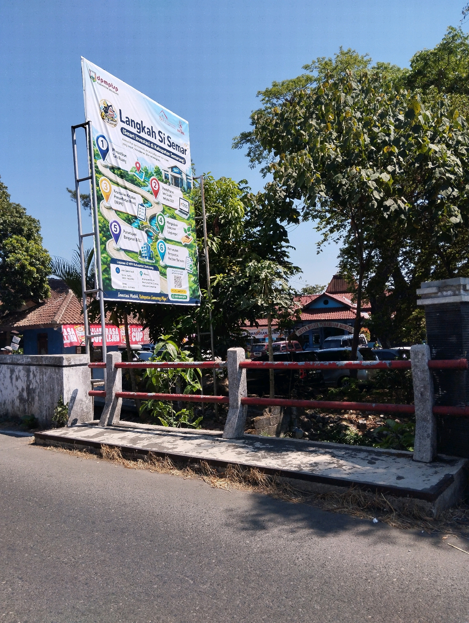 This image captures a street view in Rowoboni, Indonesia, dominated by a large white banner advertising "Langkah Si Semar (Smart Investasi di Kabupaten Semarang)". The banner is supported by metal poles and features a colorful graphic illustrating a winding path with numbered steps, each representing a service or benefit related to investment. The text on the banner is primarily in Indonesian, with phrases like "Memudahkan perizinan", "Meningkatkan daya saing", and contact information including a phone number and a QR code.
To the left of the banner, a red-tiled building with blue accents is visible, partially obscured by trees. A sign on this building reads "RASA LEJEN CUP". Further back, several cars are parked in front of another building, which has a sign that appears to say "OBYEK WISATA PEMANDIAN MUNCUL".
In the foreground, a concrete bridge with red railing spans a small waterway or ditch. The road in front of the bridge shows a textured asphalt surface. The environment is characterized by lush green trees and a clear blue sky, indicating a sunny day. The scene appears to be a public space, possibly near a government office or a tourist attraction, given the nature of the banner and the buildings. There are no people clearly visible in the immediate foreground or engaged in any specific actions.