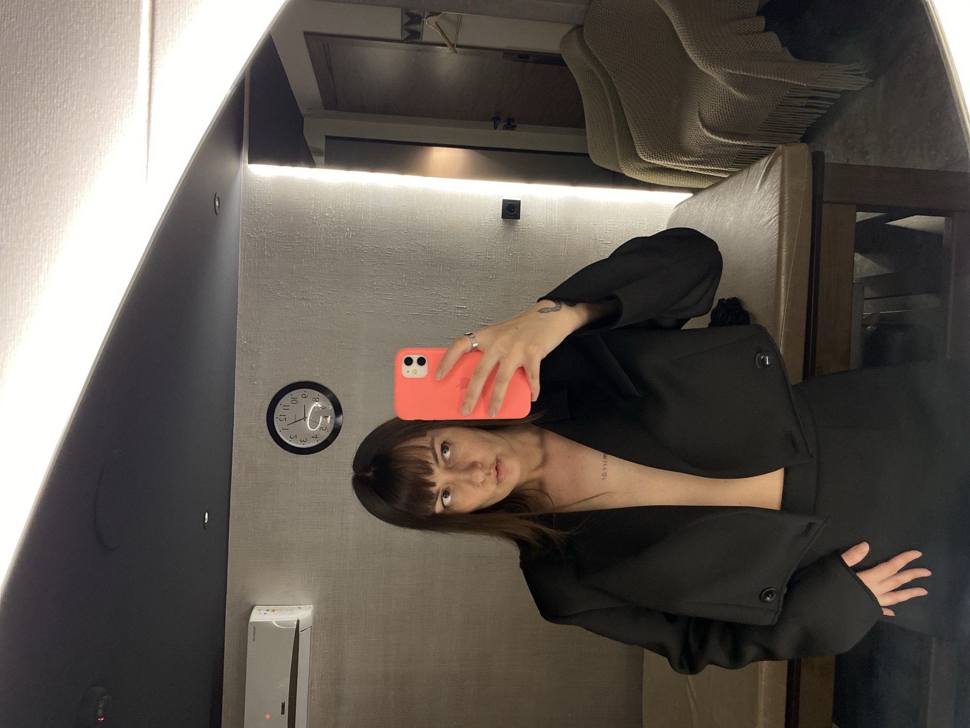 A woman is taking a selfie in a mirror. She has brown hair and is wearing a black outfit. She is holding an orange phone. Behind her, there is a clock on the wall and a bed with a blanket. The location is inside a room with light gray walls. The specific location in Votkinsk, Russia is undeterminable from the image.